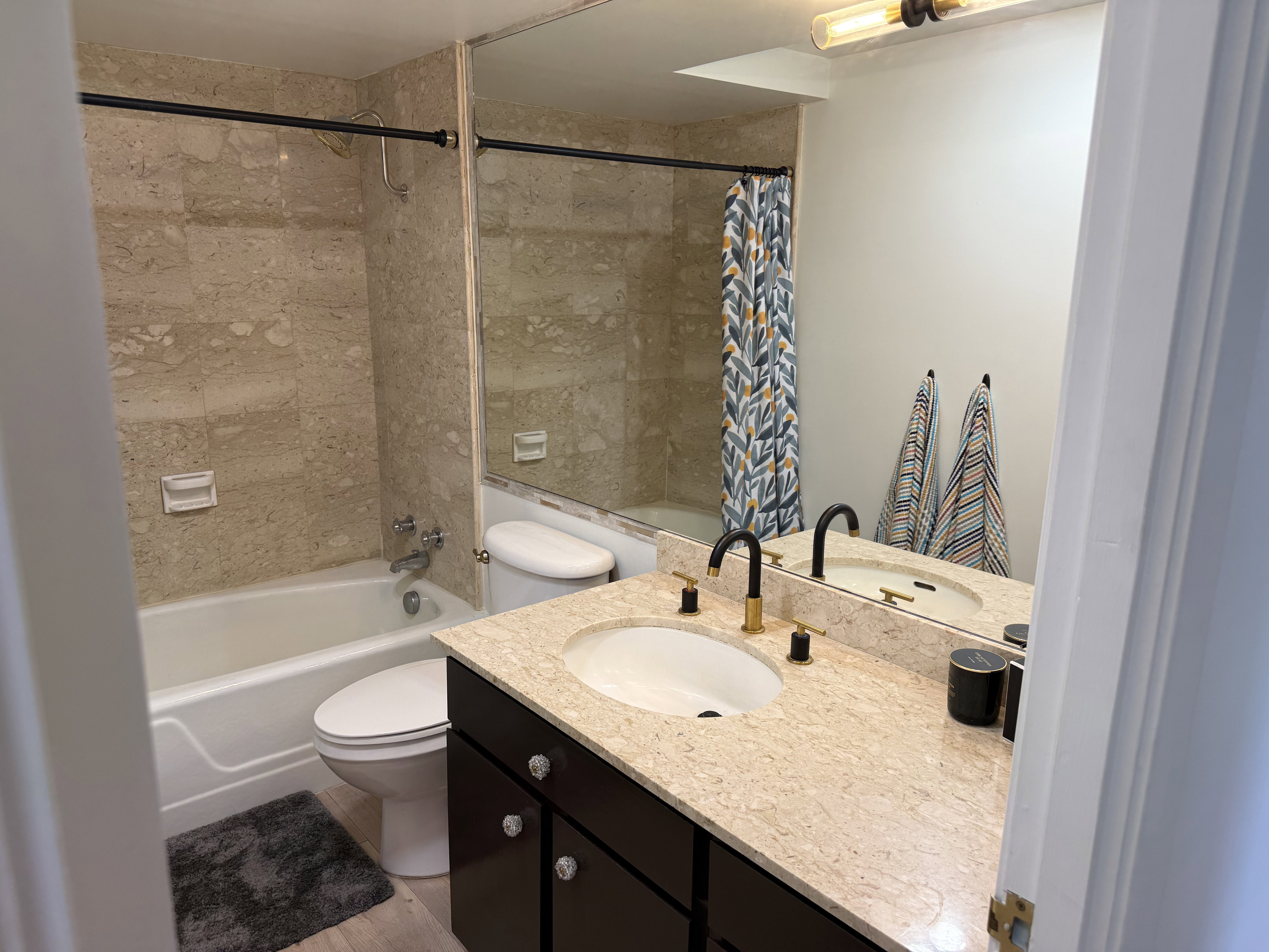 Bathroom featuring a built-in tub with tile surround, a dark wood vanity with a light-colored countertop, and a double vanity sink with black and gold fixtures