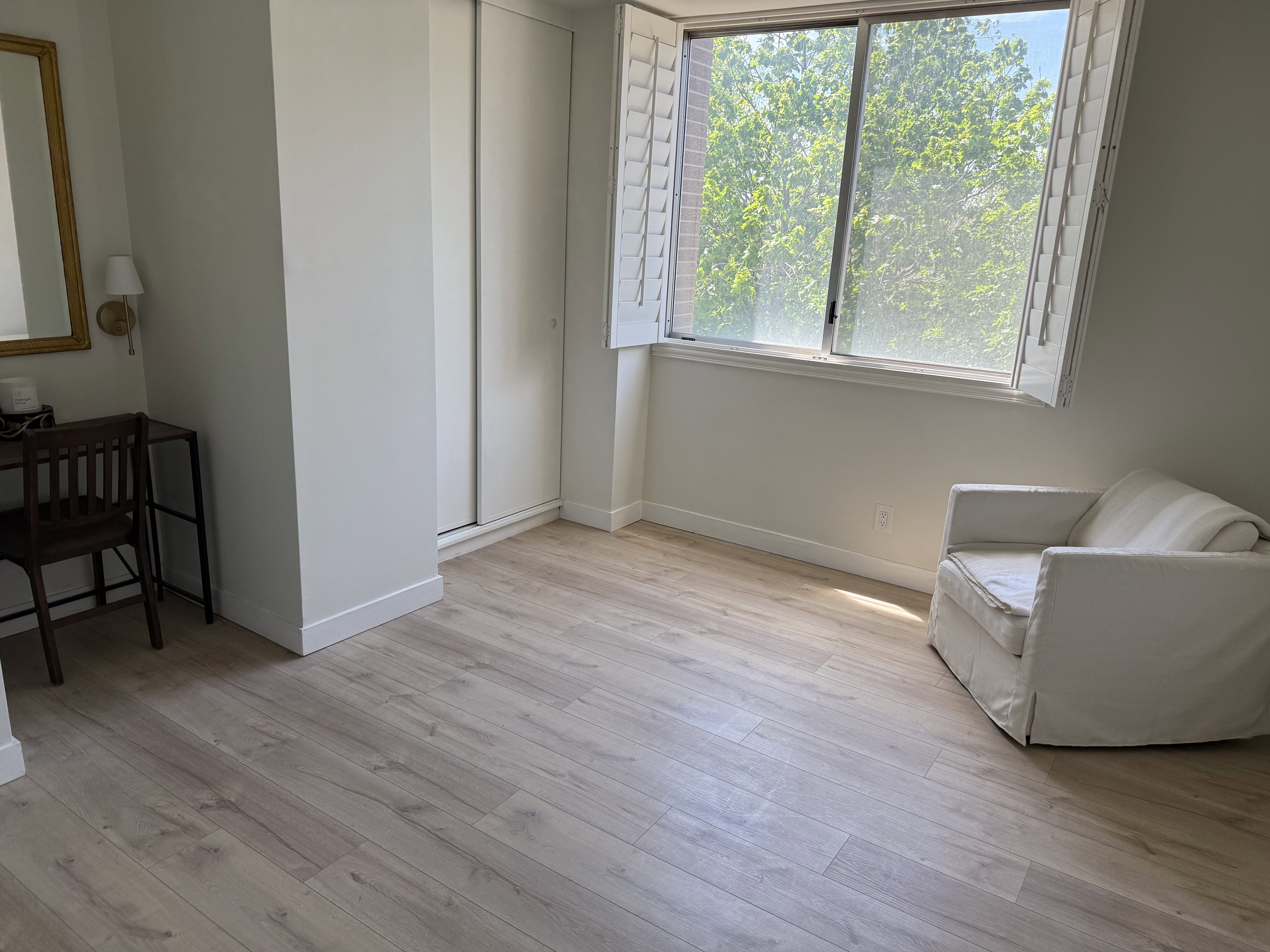 Expansive room featuring light wood-finish flooring, a large window with white plantation shutters, built-in sliding panel wardrobes, and light neutral wall paint