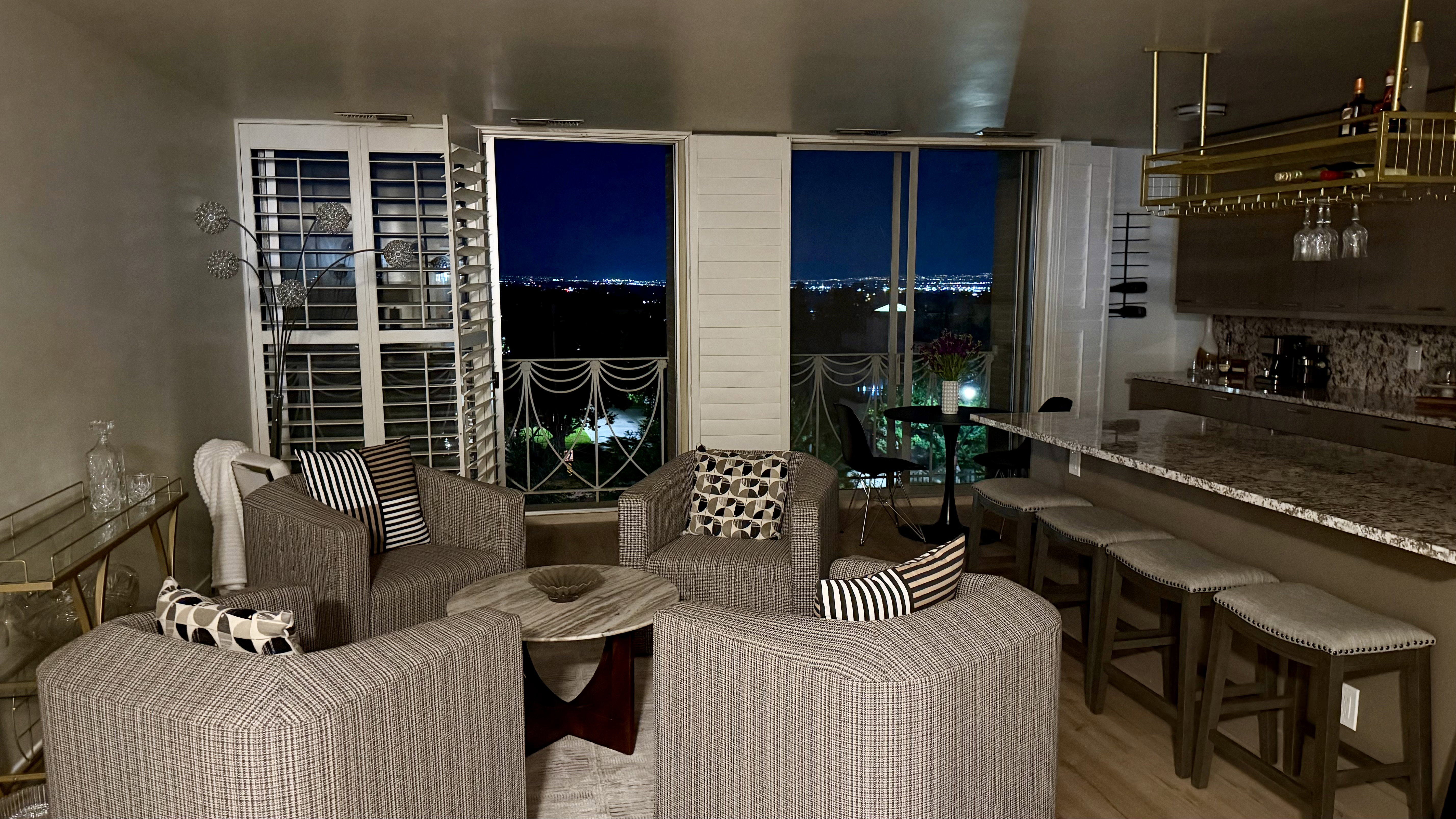 Open living area with wood-finish flooring, a built-in bar featuring a gold-finish hanging wine rack, an extended countertop with seating for four, and exterior access to a balcony through sliding glass doors