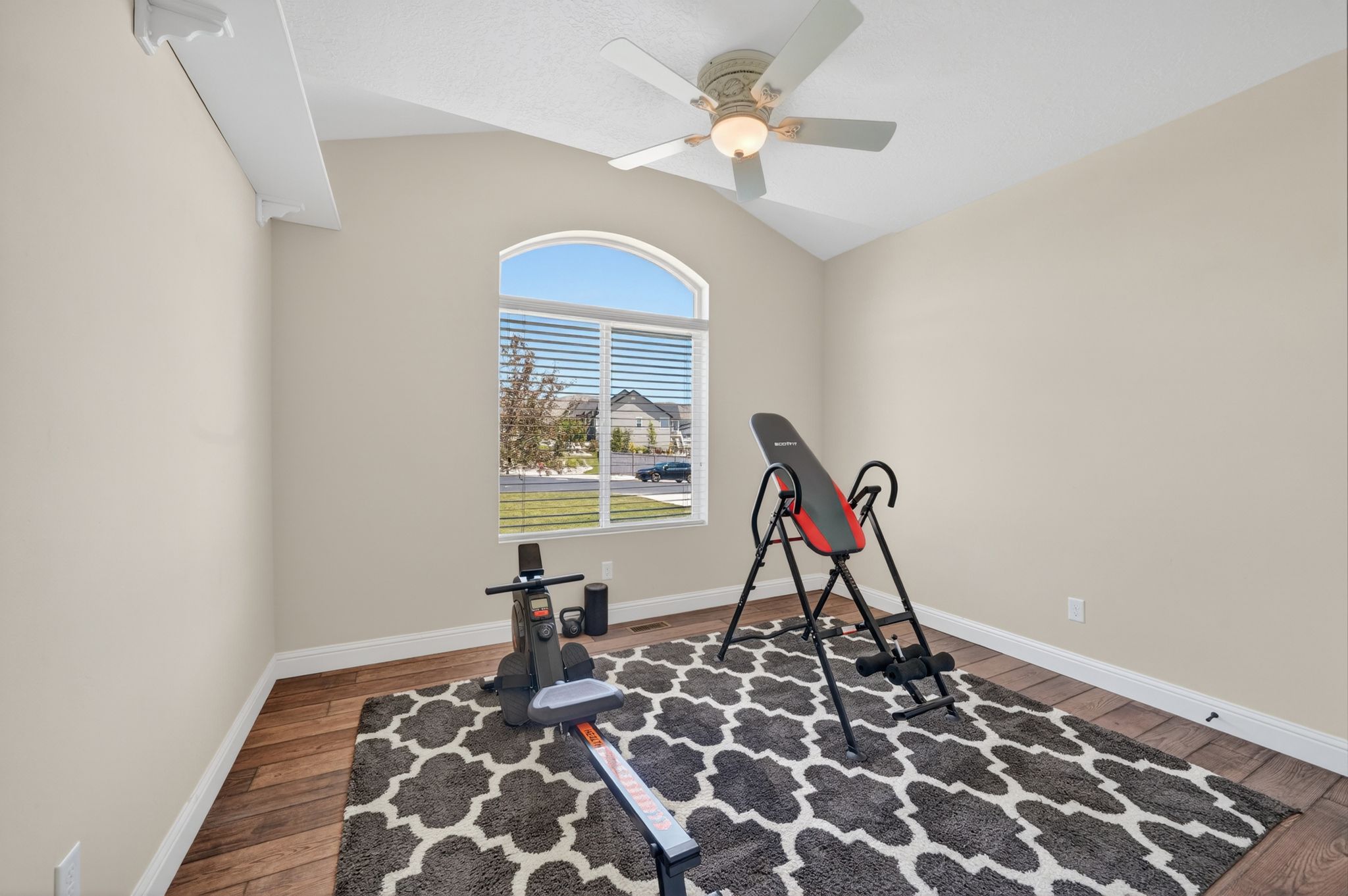 Vaulted ceiling room featuring a large arched window with horizontal blinds, wood-finish flooring, and a ceiling fan with light fixture