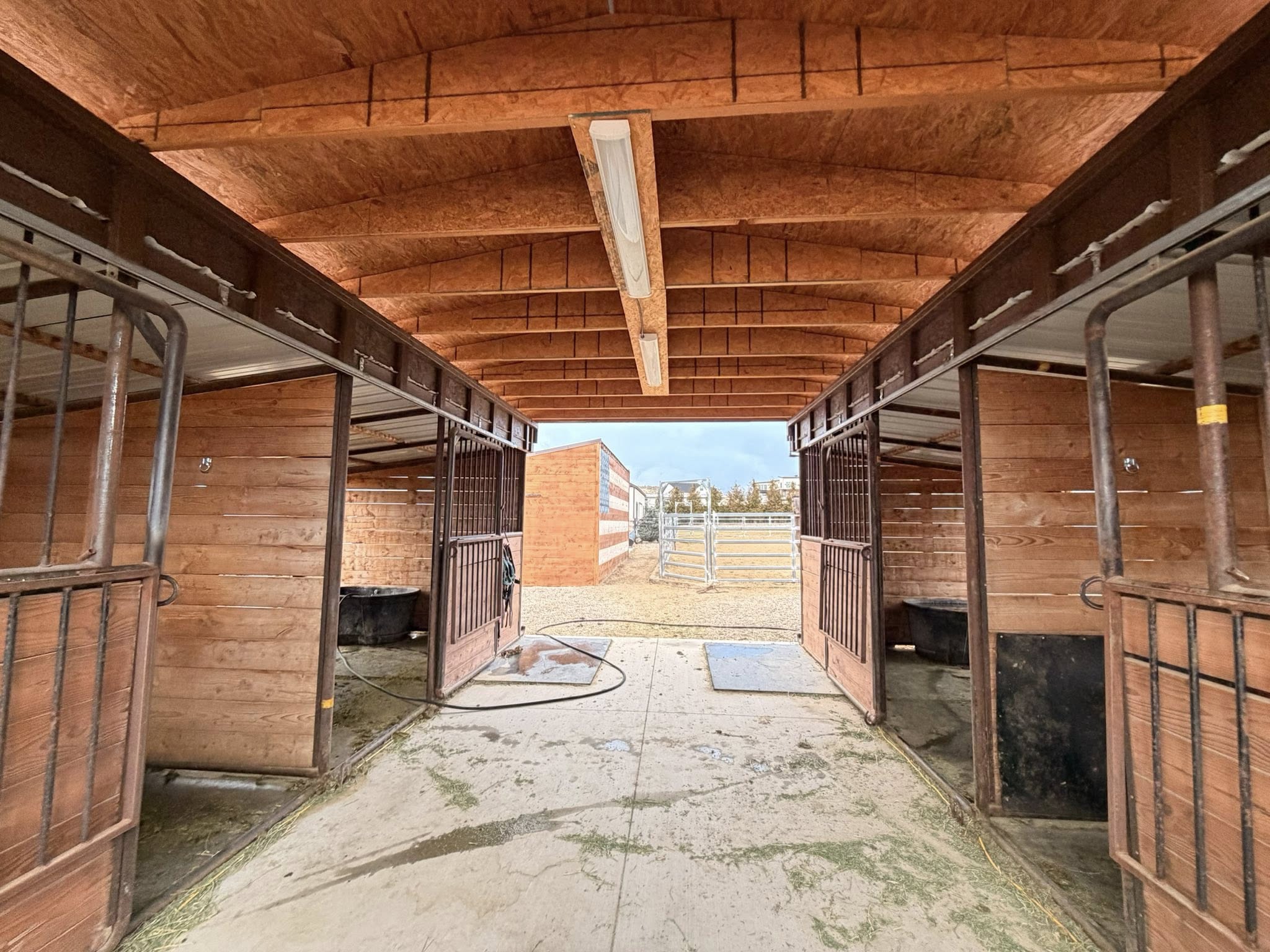 Covered equestrian stalls featuring wood paneling, metal gates, and an exposed wood ceiling with fluorescent lighting