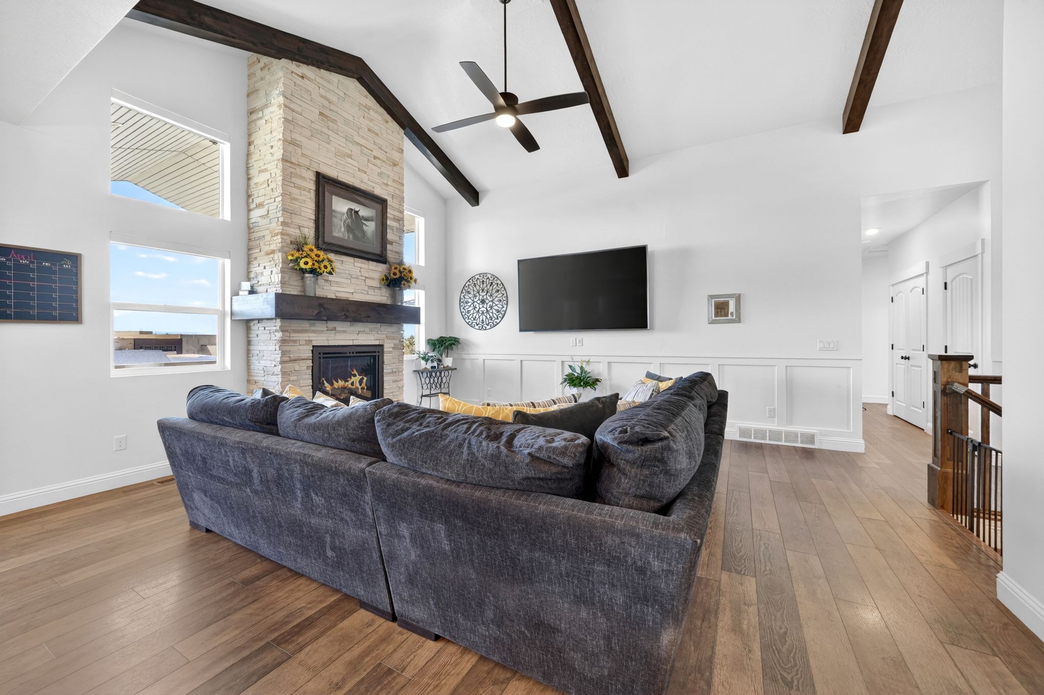 Vaulted ceiling with exposed wood beams, stone-clad fireplace with wood mantle, multiple windows, wood-finish flooring, and wainscoting