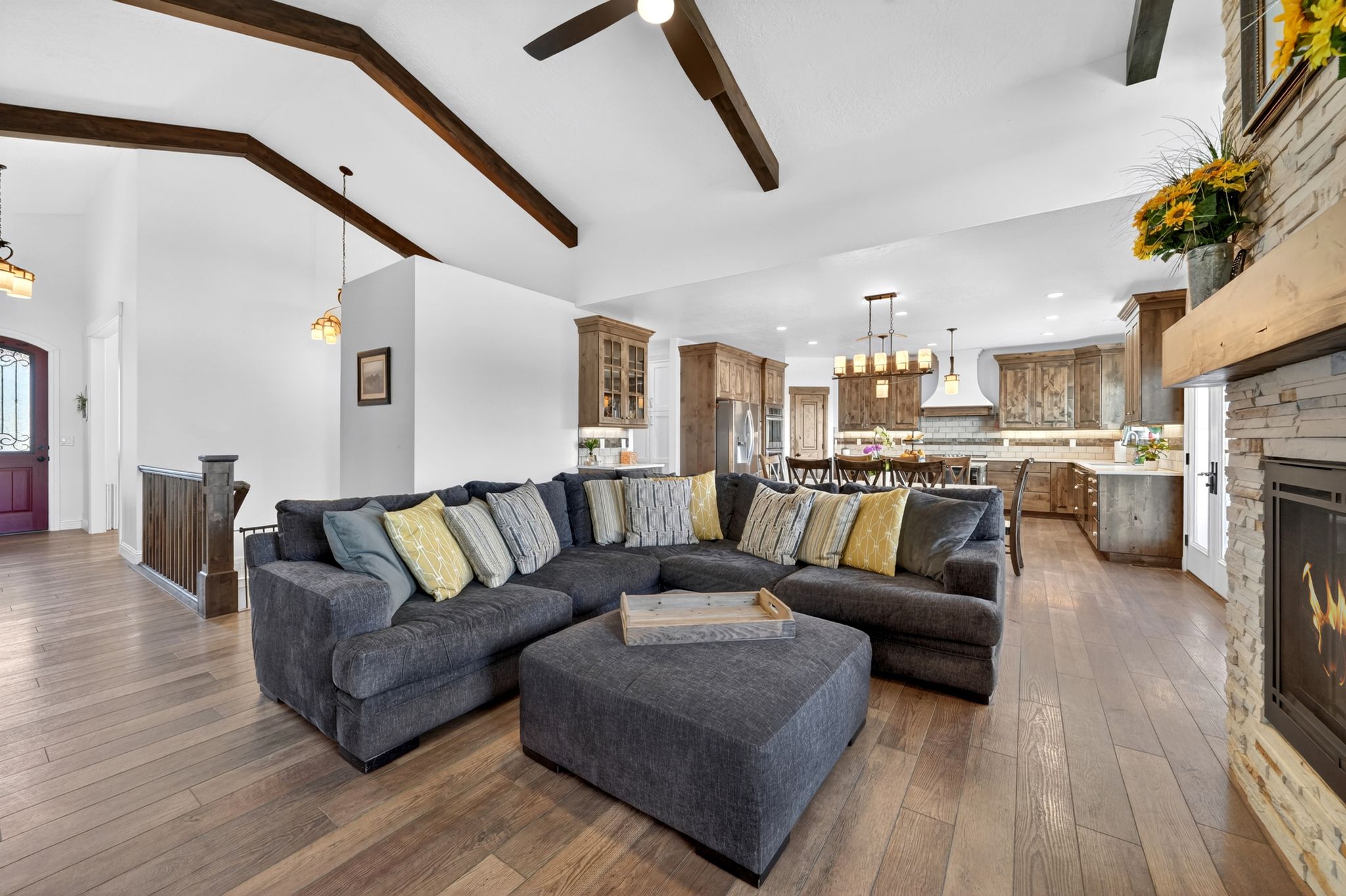 Spacious open-concept living area featuring wood-finish flooring, vaulted ceilings with exposed wood beams, and a stone-clad fireplace with a wood mantle