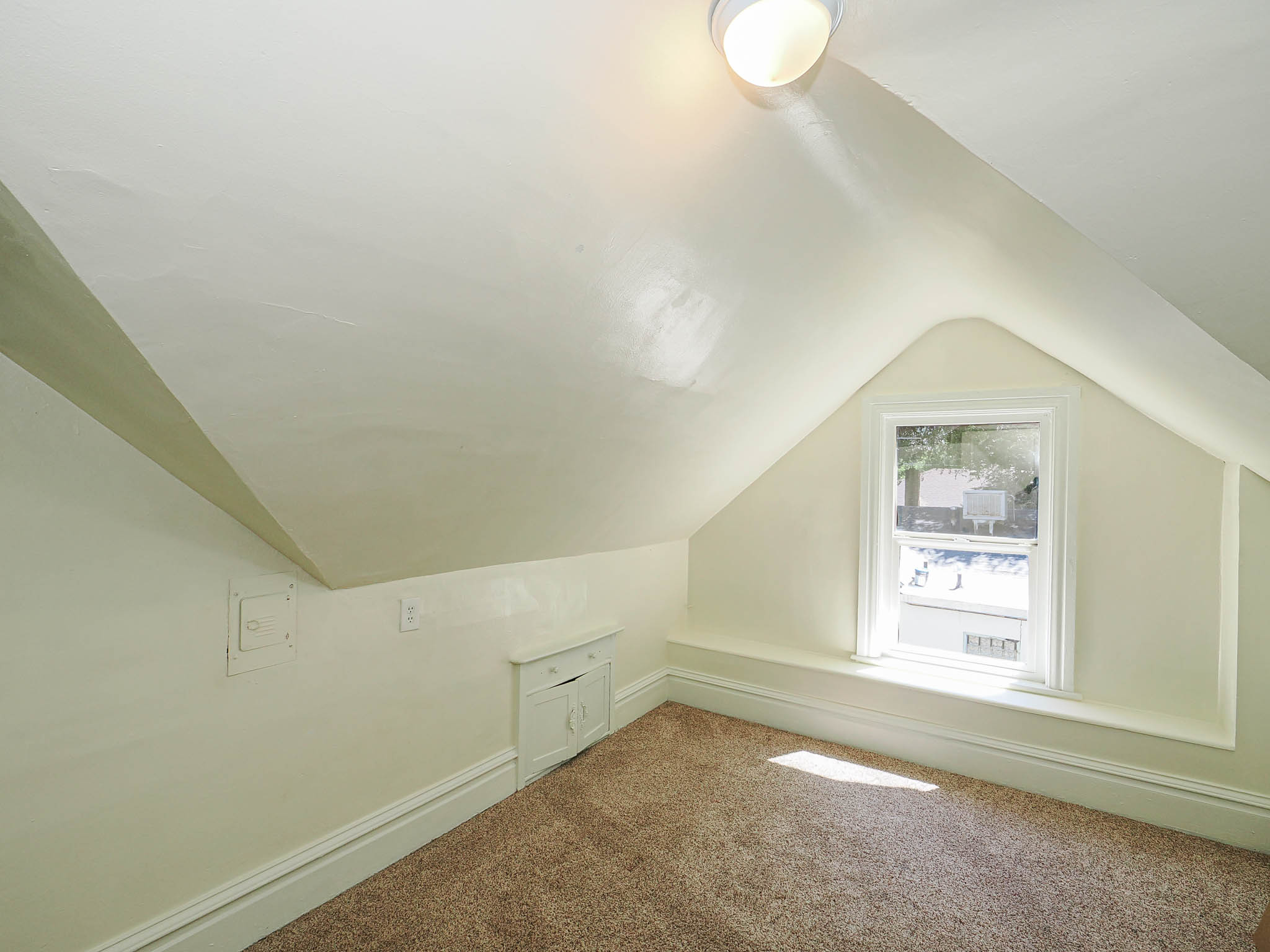 Attic space featuring a single window, sloped ceilings, wall-mounted light fixture, and brown carpeting