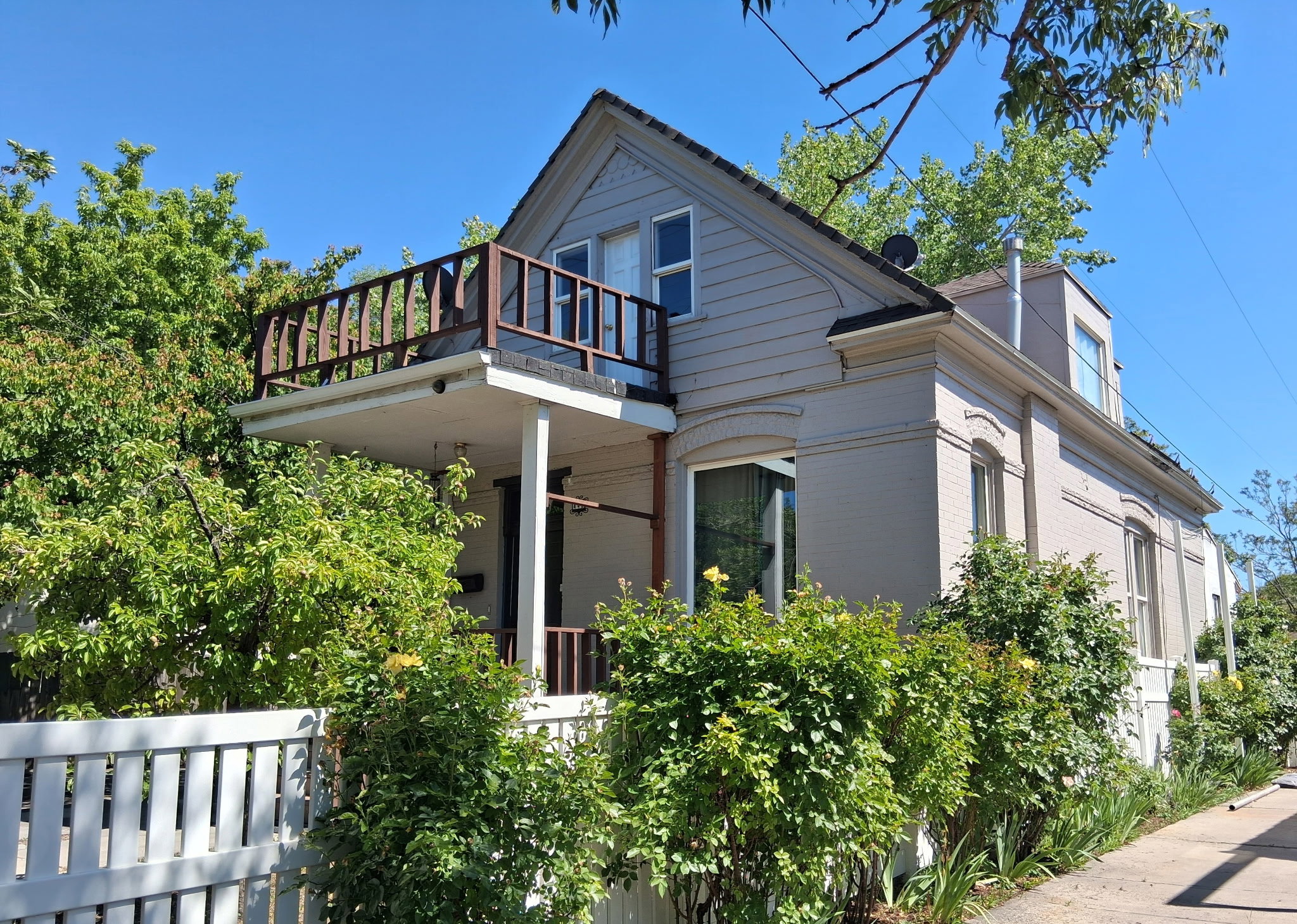 Exterior featuring a white picket fence, mature landscaping, second-story balcony with wood railing, and dormer window