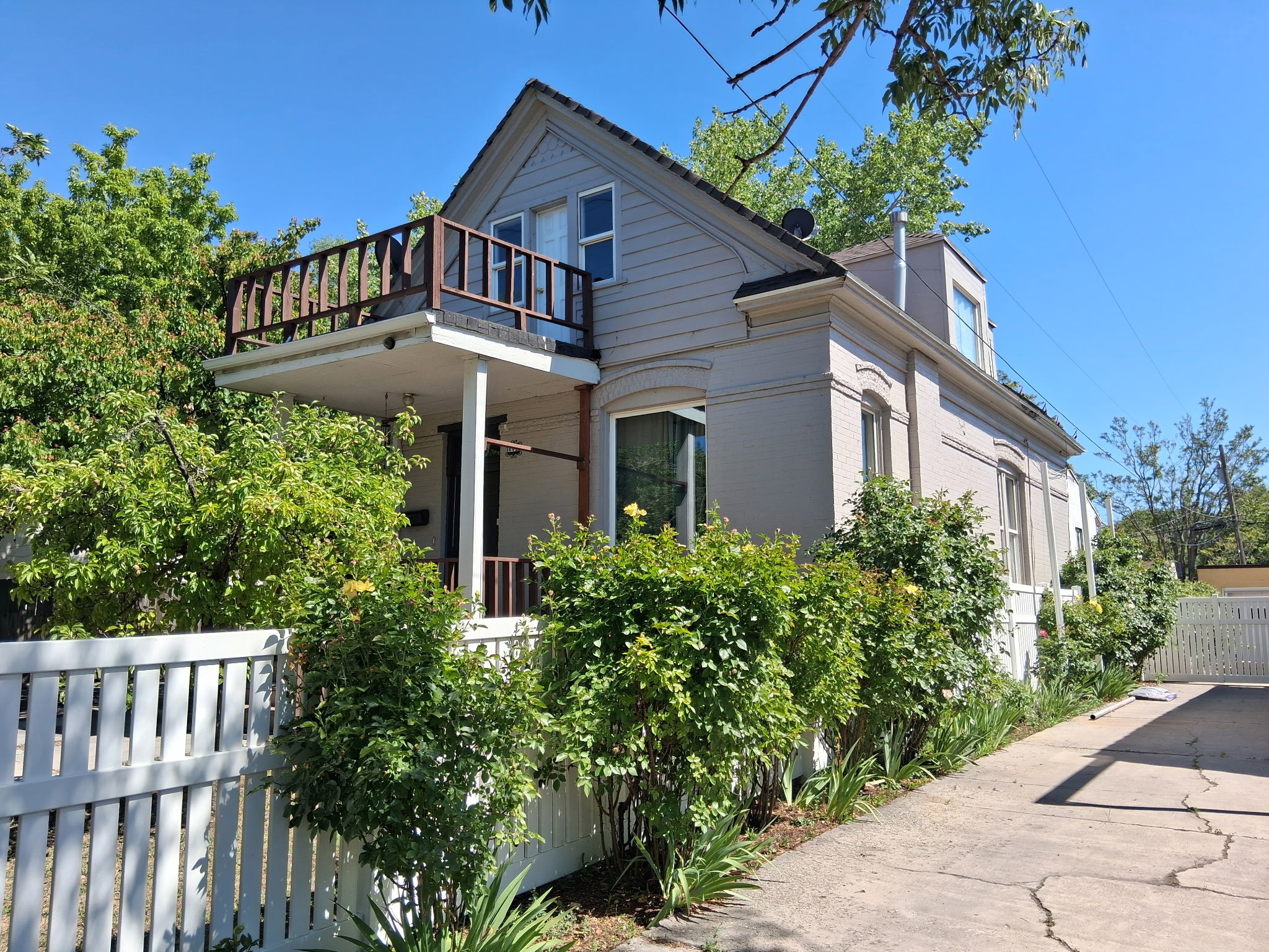 Two-story residence featuring a second-level balcony with wood railing, a white picket fence, mature perimeter shrubs, and a concrete driveway