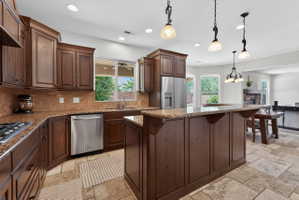 Spacious kitchen featuring dark wood cabinetry, stainless steel appliances, a large island with an extended countertop, stone tile flooring, and recessed lighting