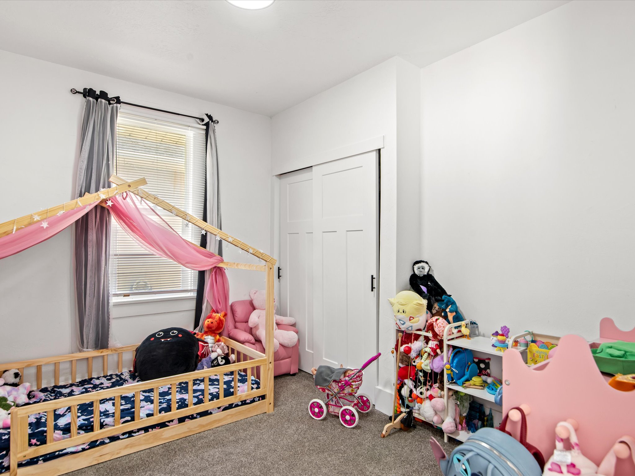 Bright room featuring a window with horizontal blinds, neutral carpet, white walls, a white ceiling light fixture, and a white sliding closet door with dark hardware