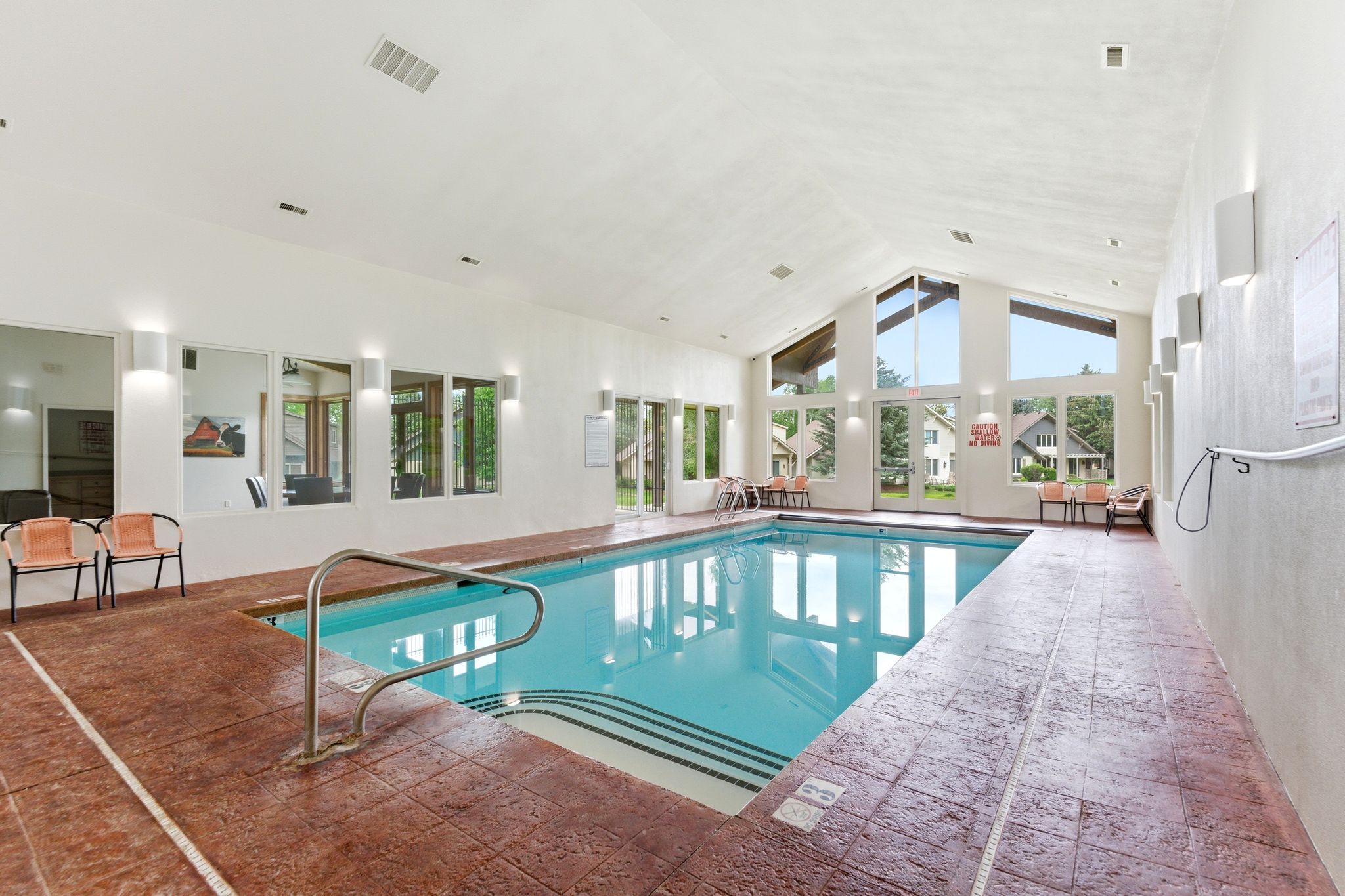 Indoor swimming pool featuring a vaulted ceiling with exposed beams, expansive windows, stamped concrete decking, and multiple wall sconces
