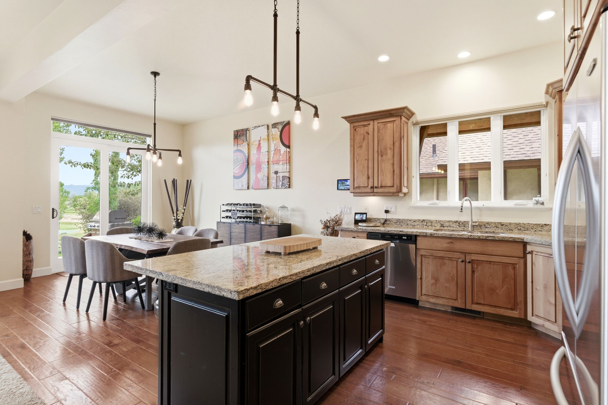Open-concept kitchen and dining area featuring wood-finish flooring, a dark kitchen island with a light-toned countertop, a kitchen perimeter with wood cabinetry and a light-toned countertop, and a dining area with a glass slider