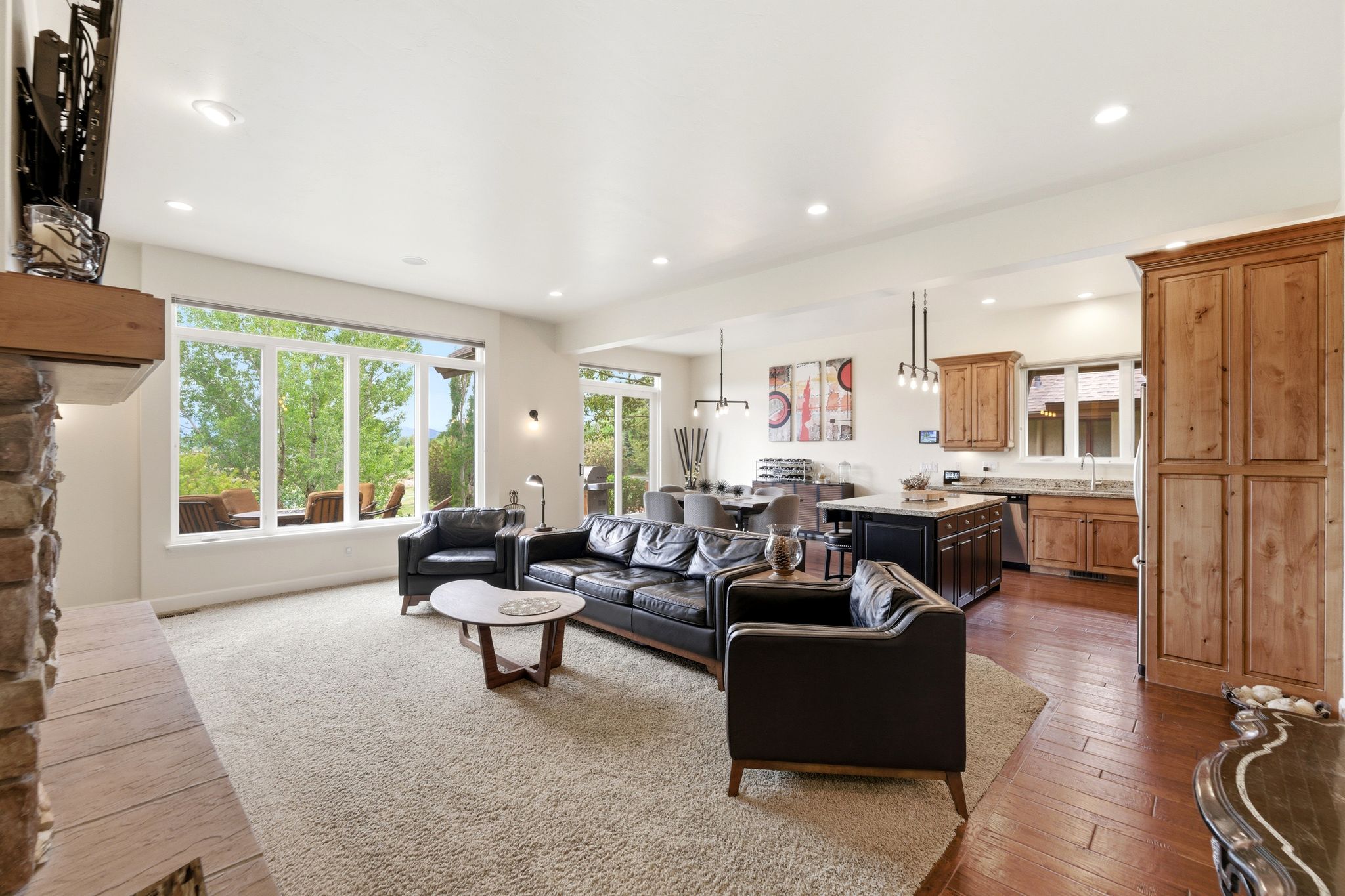 Open-concept living space featuring a stone fireplace with a wood mantle, a wall of windows, recessed lighting, and wood-finish flooring in the kitchen and dining areas