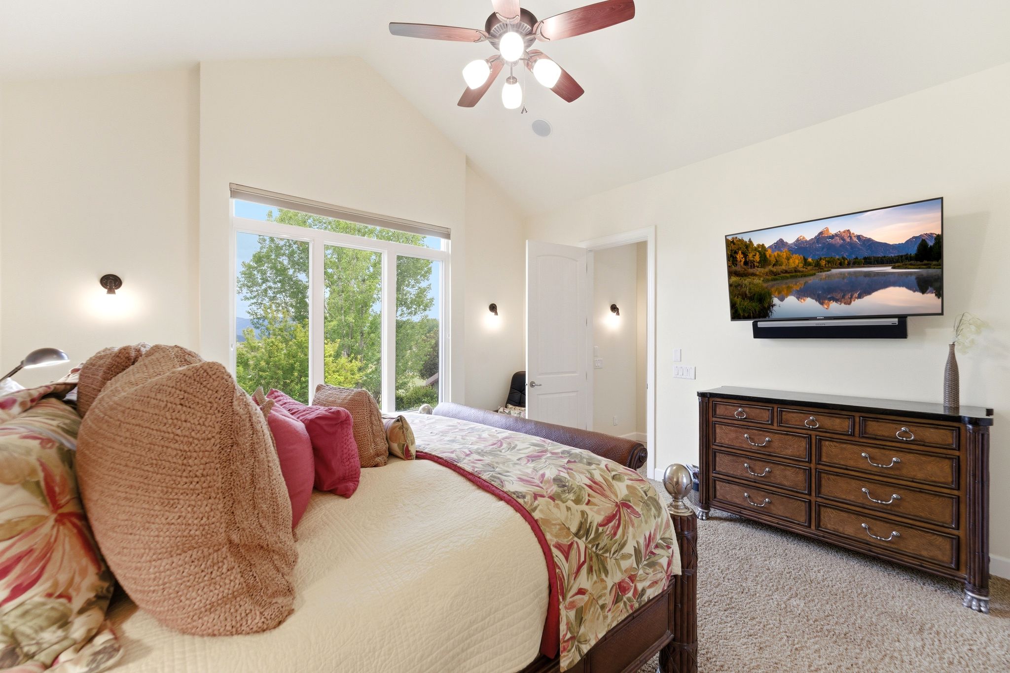 Vaulted ceiling bedroom with a large picture window, a ceiling fan, and two wall-mounted light fixtures