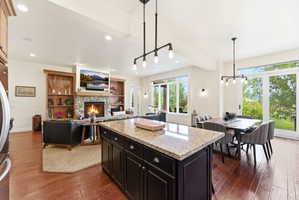 Open-concept living space featuring a central kitchen island with granite countertop, a stone fireplace with a wood mantle and built-in shelving, wood-finish flooring, a dining area with pendant lighting, and large glass sliding doors providing exterior a