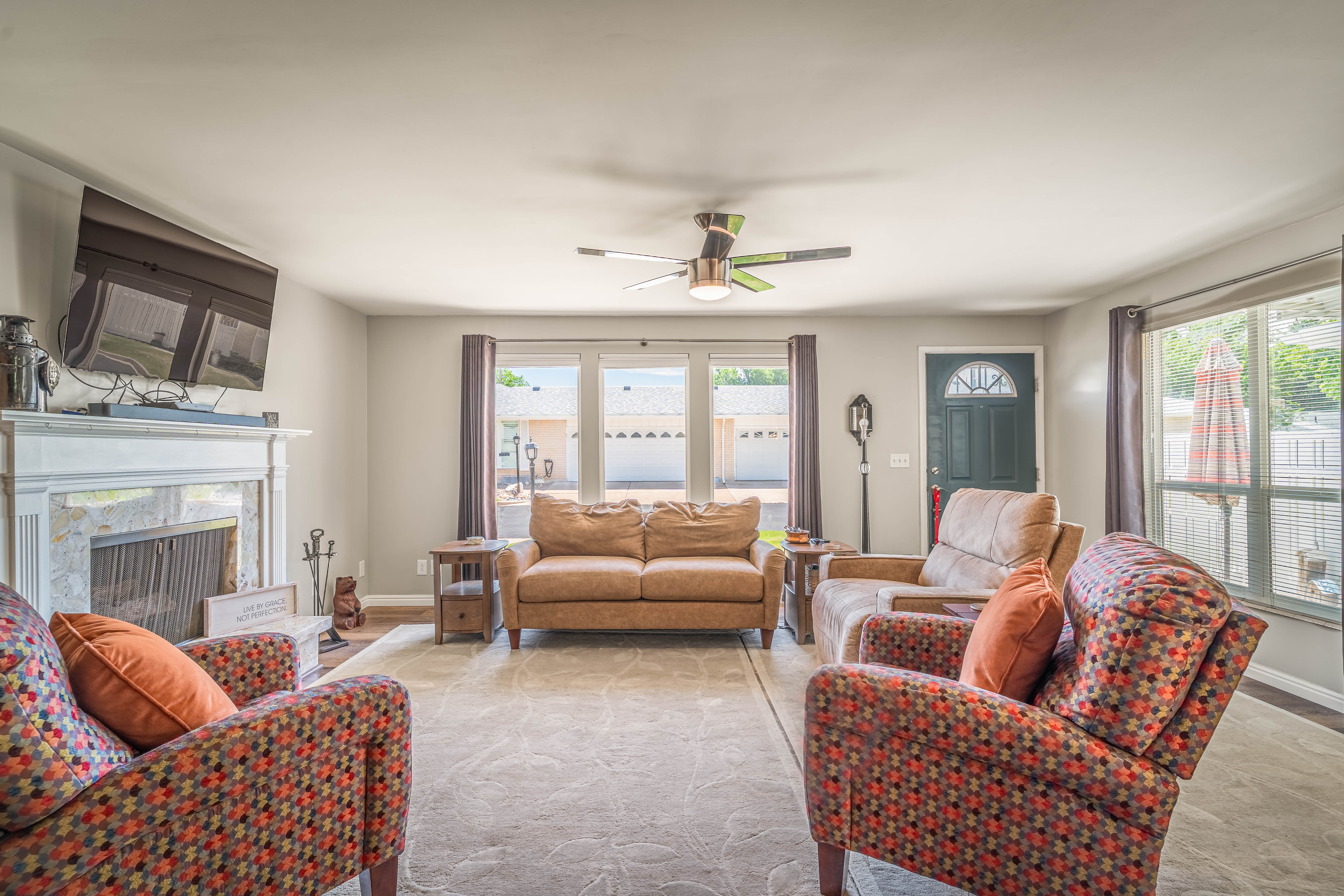Living area featuring a white mantel fireplace with tile surround, a dark wood-finish front entry door with decorative glass insert, a ceiling fan with integrated lighting, and multiple windows
