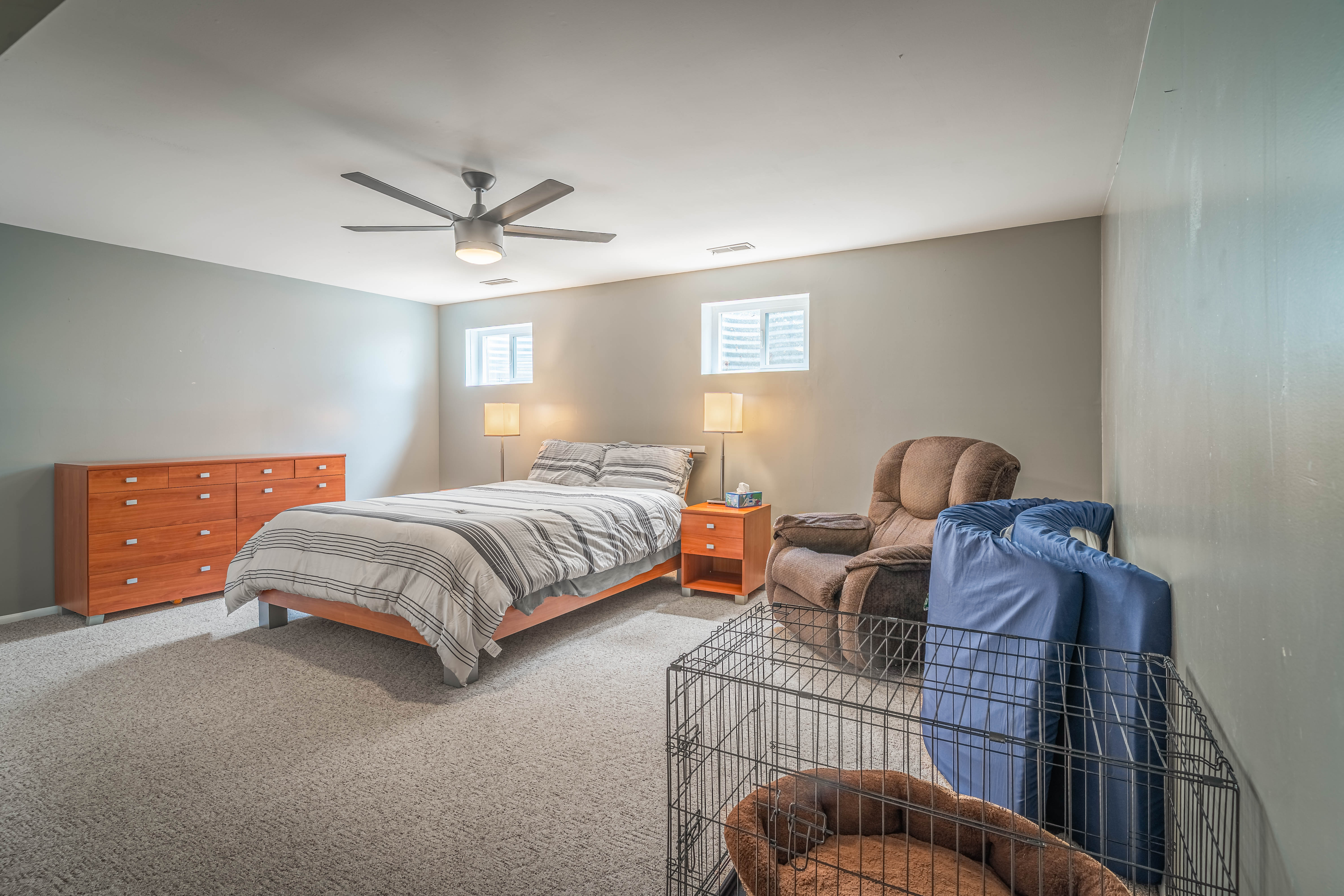 Spacious room featuring neutral carpeting, light gray walls, a ceiling fan with integrated lighting, and two windows with white louvered blinds