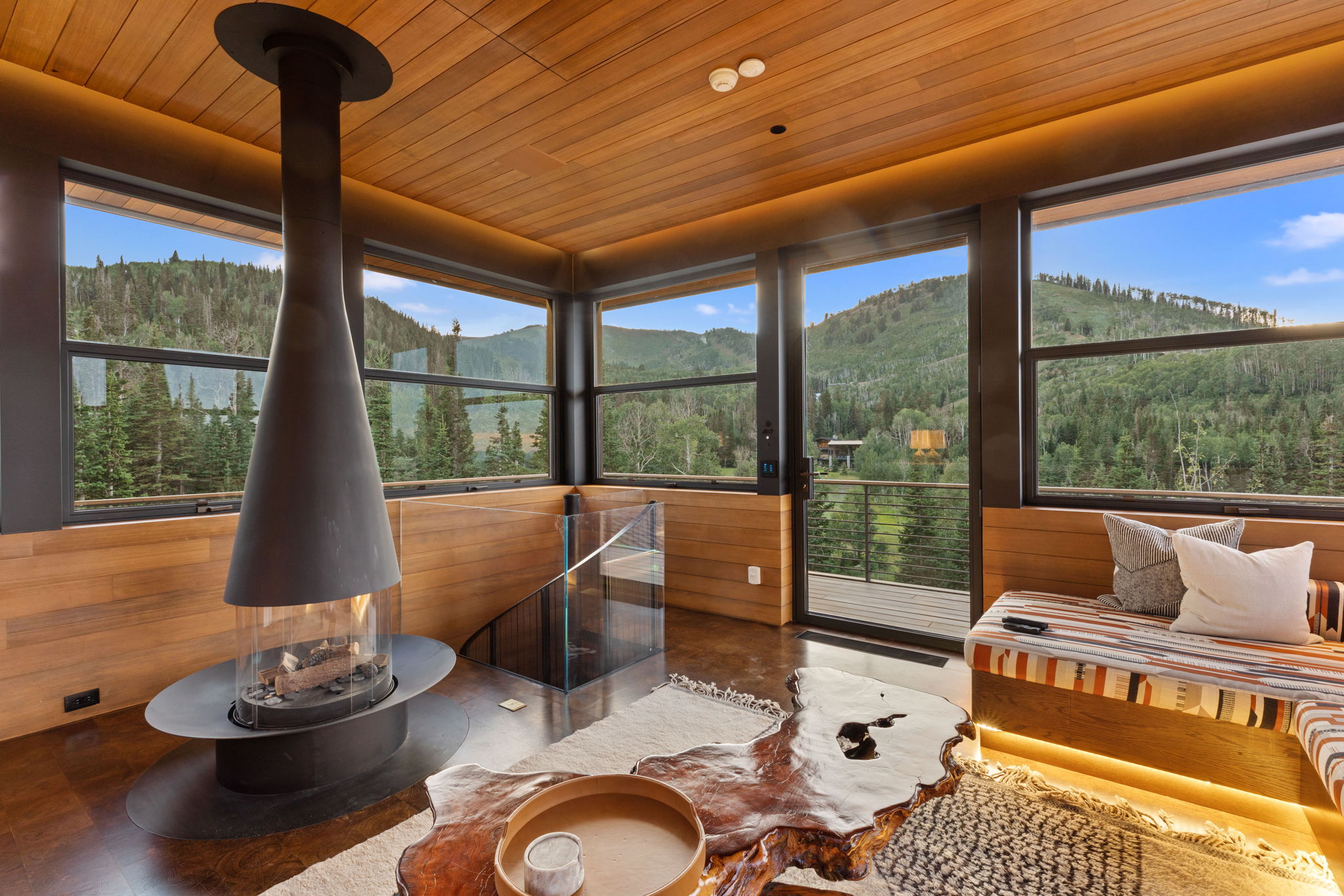 Modern freestanding fireplace with a glass enclosure, wood-finish flooring, full-height windows, wood paneled walls, and a built-in seating area