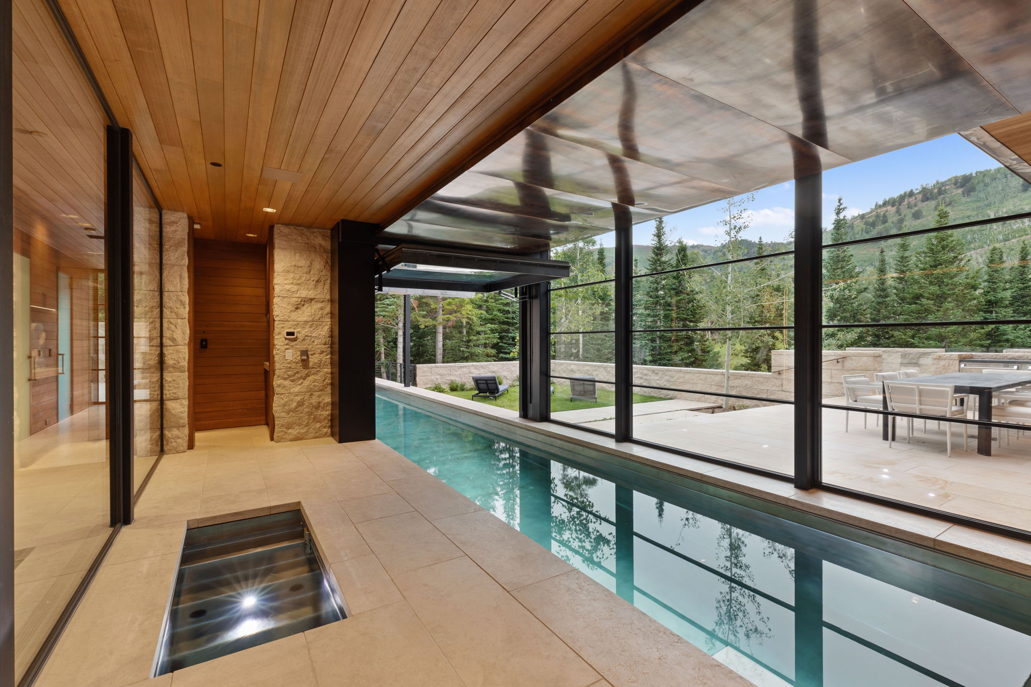 Indoor lap pool featuring a retractable glass wall, wood-paneled ceiling, stone accent walls, and a large outdoor patio with mountain scenery
