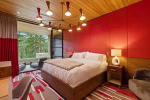 Floor-to-ceiling glass doors opening onto an exterior stone patio with seating, wood plank ceiling, accent wall with red upholstered panels, and a patterned area rug