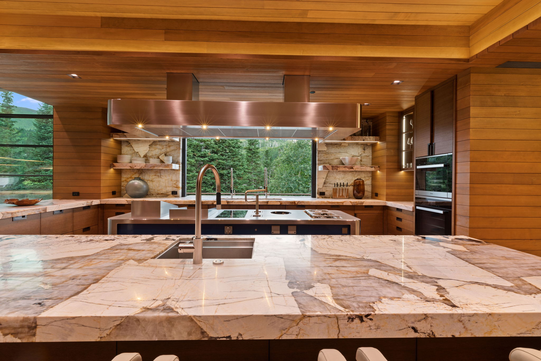 Contemporary kitchen featuring extensive natural wood paneling, double islands with stone countertops, a stainless steel range hood, built-in double ovens, and a large window providing an expansive outdoor scene