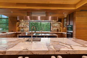 Contemporary kitchen featuring extensive natural wood paneling, double islands with stone countertops, a stainless steel range hood, built-in double ovens, and a large window providing an expansive outdoor scene
