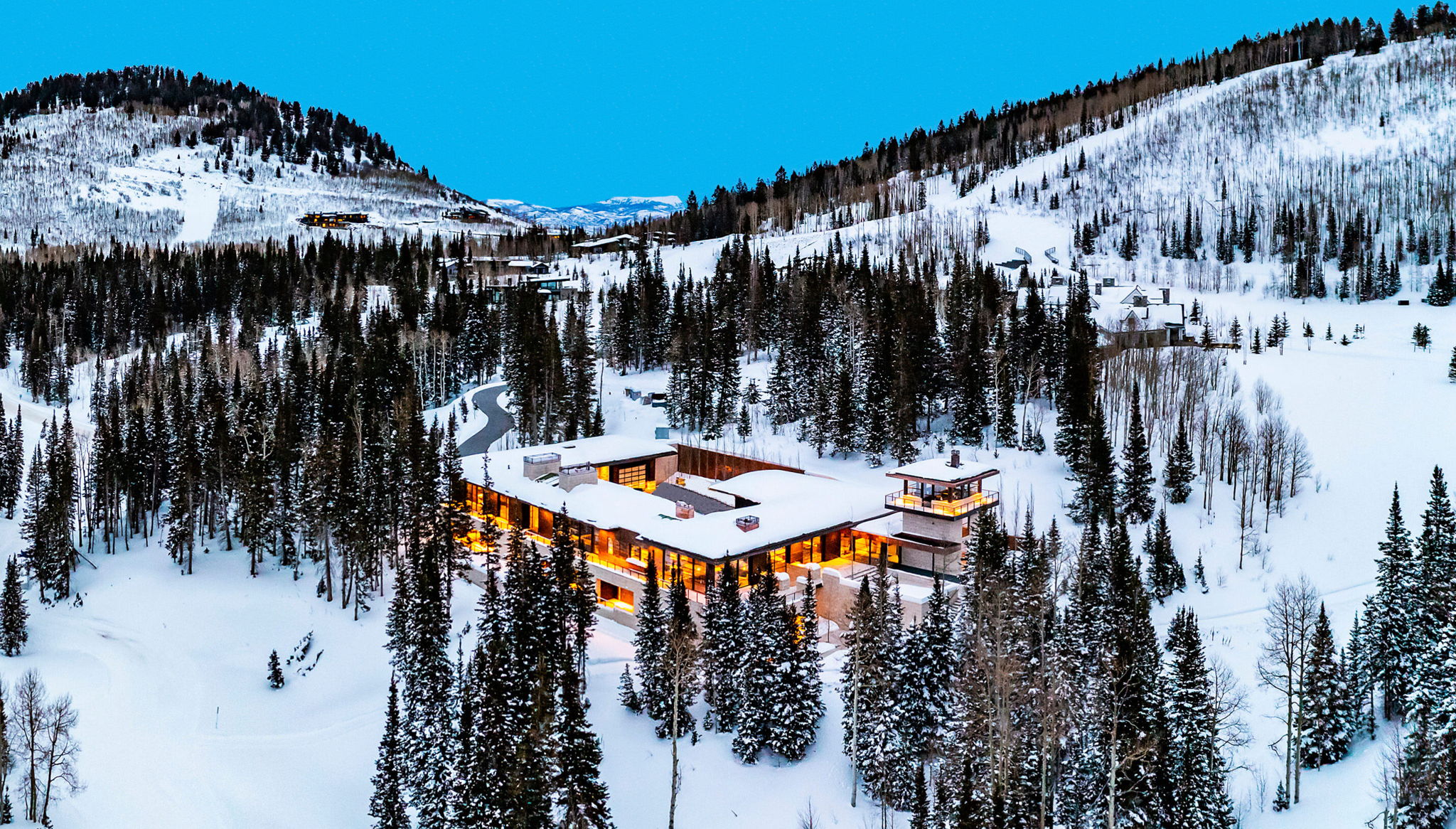 Expansive mountain property featuring a large residence with multiple levels, extensive exterior lighting, a prominent tower element, and surrounding evergreen trees