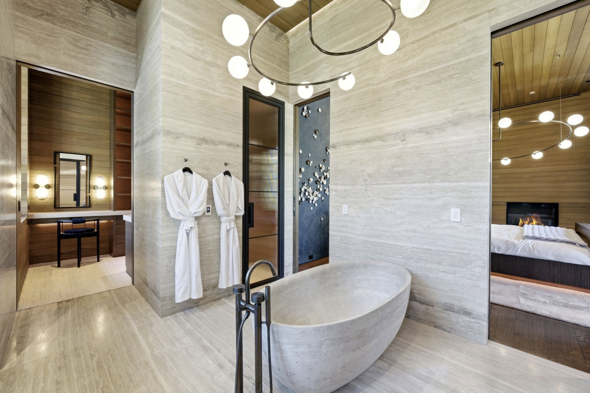 Spacious bathroom featuring a freestanding stone soaking tub, light-toned stone-finish flooring, a modern chandelier, and access to a separate vanity area with built-in shelving