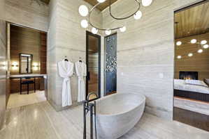 Spacious bathroom featuring a freestanding stone soaking tub, light-toned stone-finish flooring, a modern chandelier, and access to a separate vanity area with built-in shelving