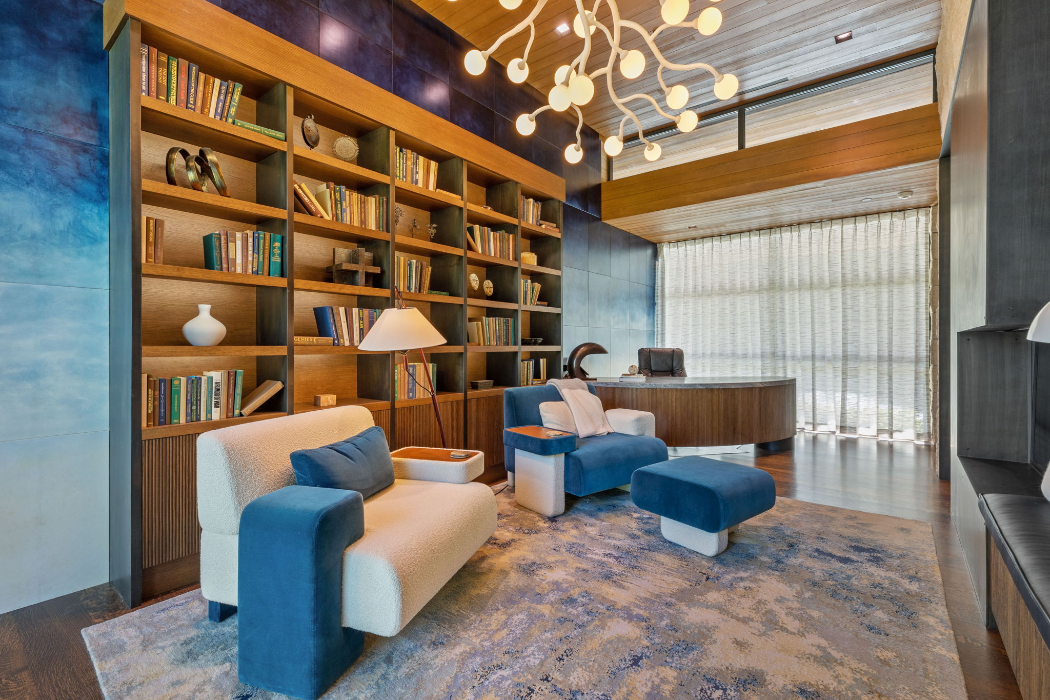 Built-in wood shelving with extensive storage, wood-finish flooring, a contemporary ceiling fixture, a curved wood desk, and a large window with privacy coverings