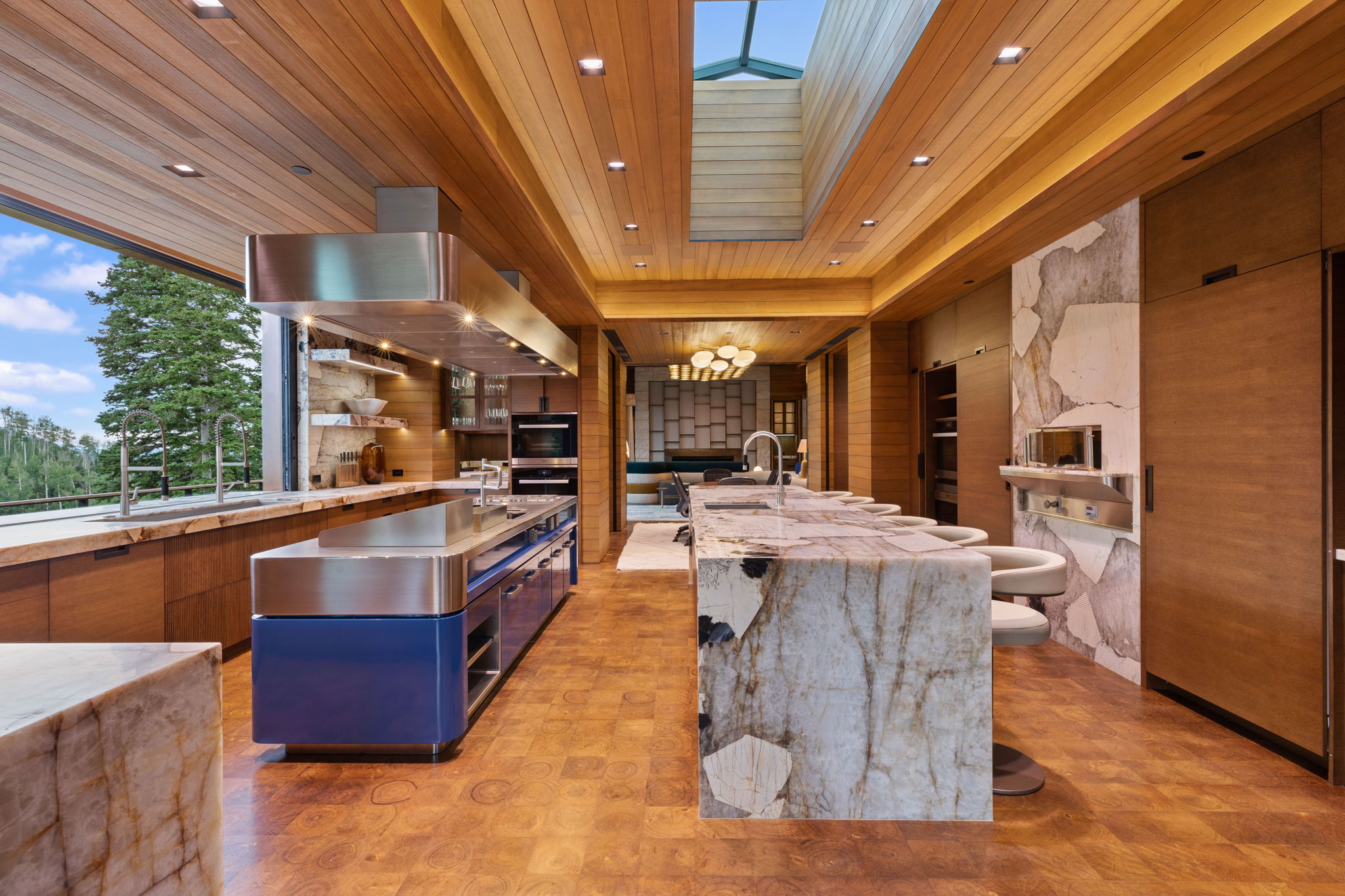 Contemporary kitchen featuring wood-finish flooring and ceilings, an expansive skylight, a large kitchen island with integrated bar seating, a second island with an integrated cooktop and stainless steel hood, and built-in appliances