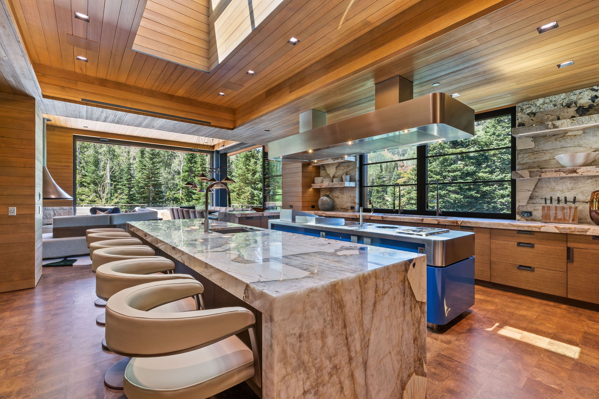 Expansive kitchen featuring a large stone slab island with seating, integrated stainless steel range hood, full-height windows, wood-finish flooring, and natural stone accent walls