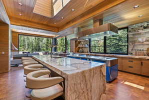 Expansive kitchen featuring a large stone slab island with seating, integrated stainless steel range hood, full-height windows, wood-finish flooring, and natural stone accent walls