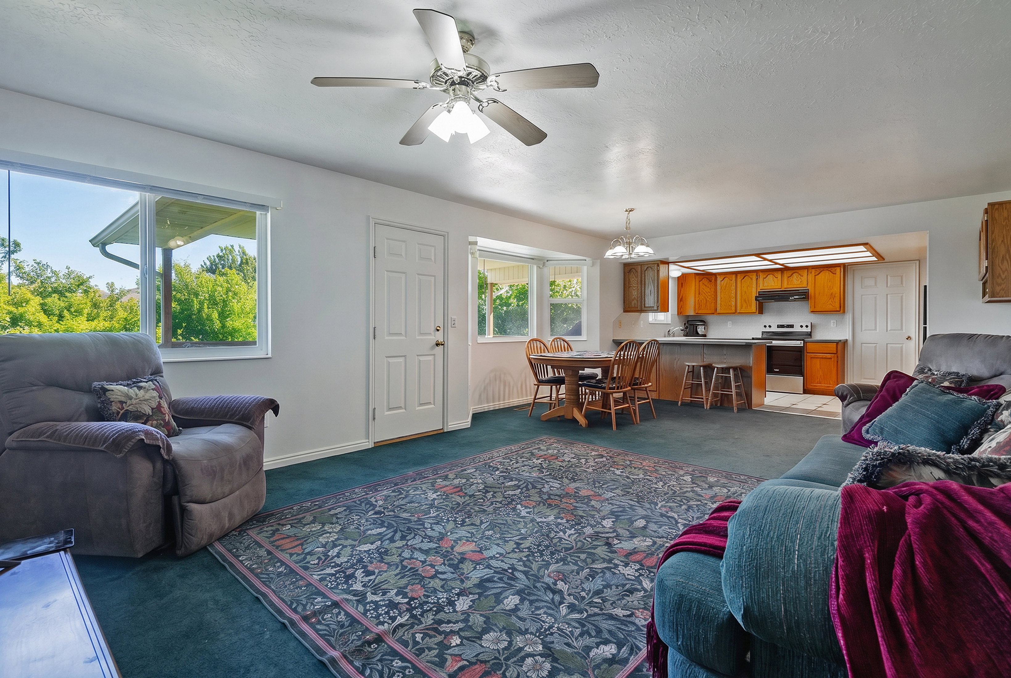 Open-concept living area featuring a ceiling fan, broad windows, and a carpeted floor