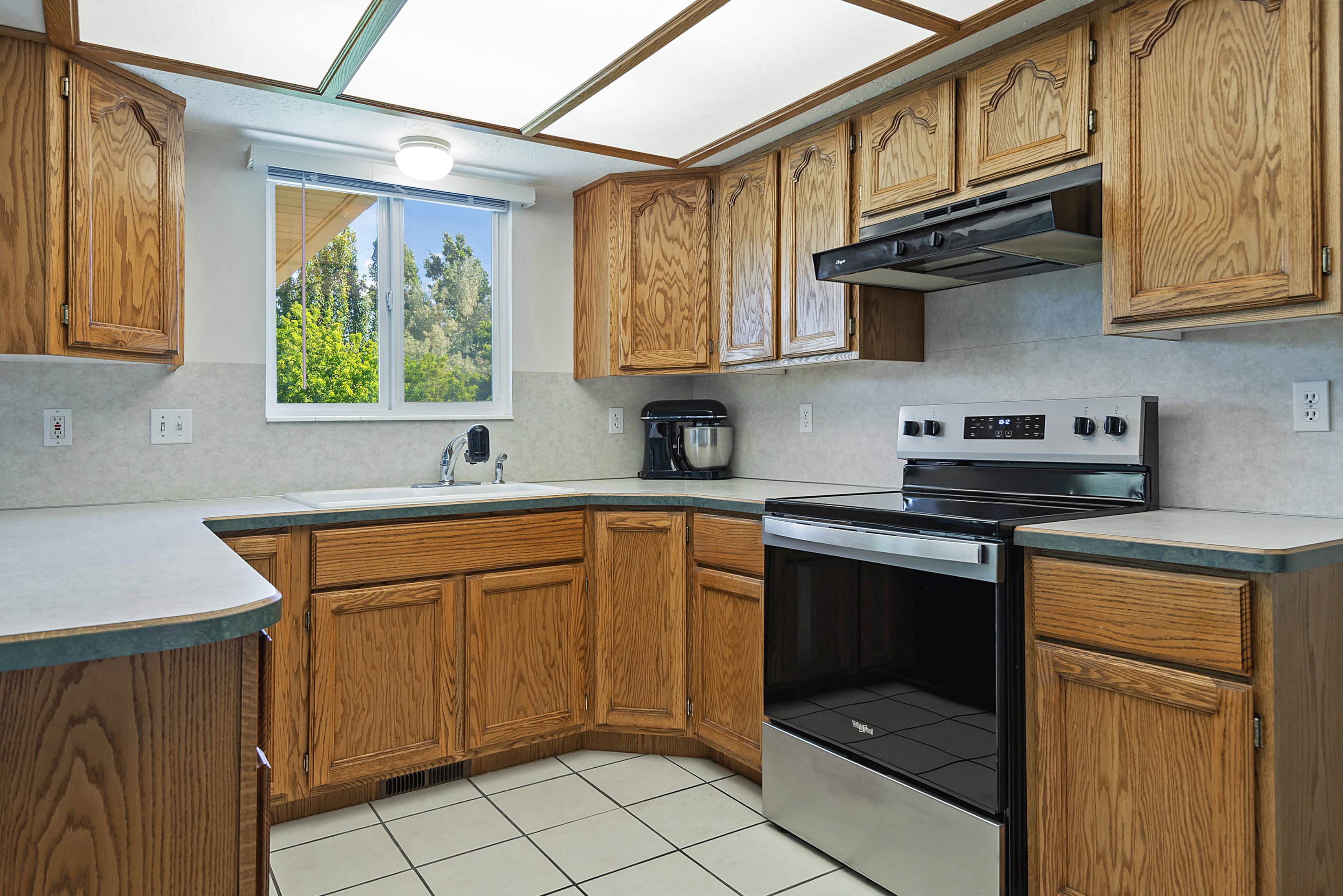 Kitchen featuring wood cabinetry, white tile flooring, stainless steel electric range, and a window overlooking exterior greenery