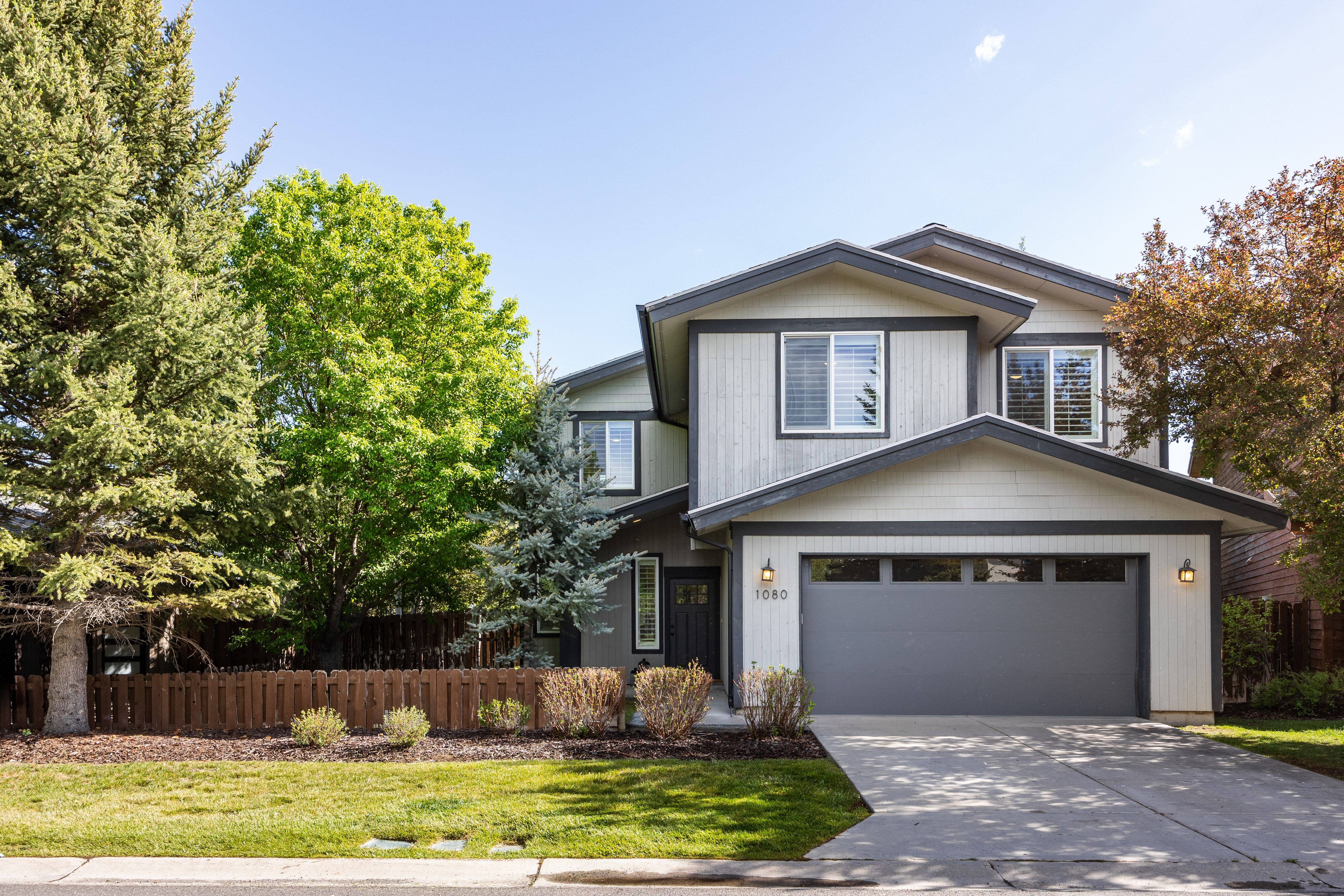 Contemporary two-story residence featuring light-toned horizontal siding with dark trim, an attached two-car garage with transom windows, a concrete driveway, and mature trees providing natural landscaping