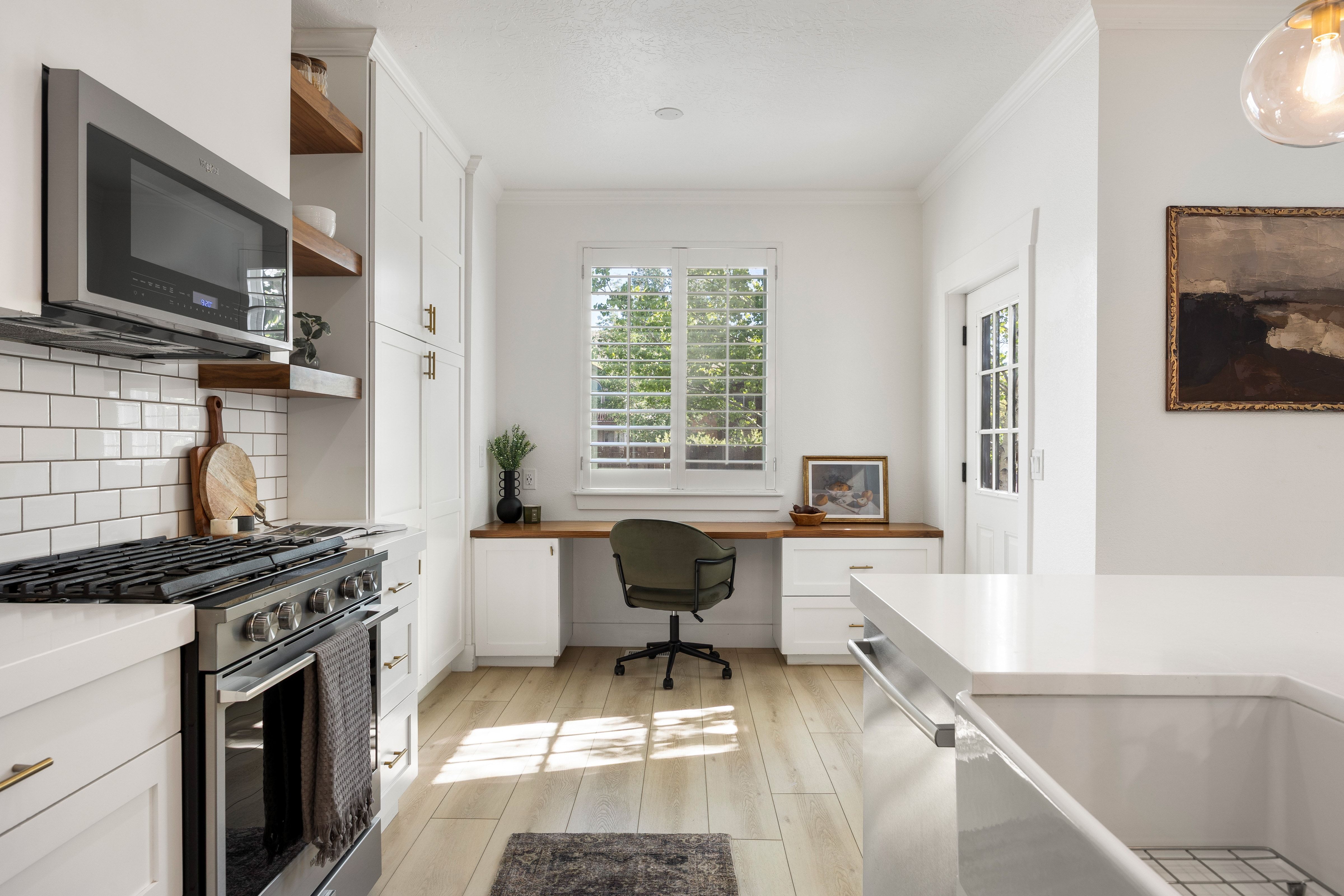 Bright kitchen workspace featuring white cabinetry, a wood-finish desk, a window with plantation shutters, a stainless steel range, and a subway tile backsplash