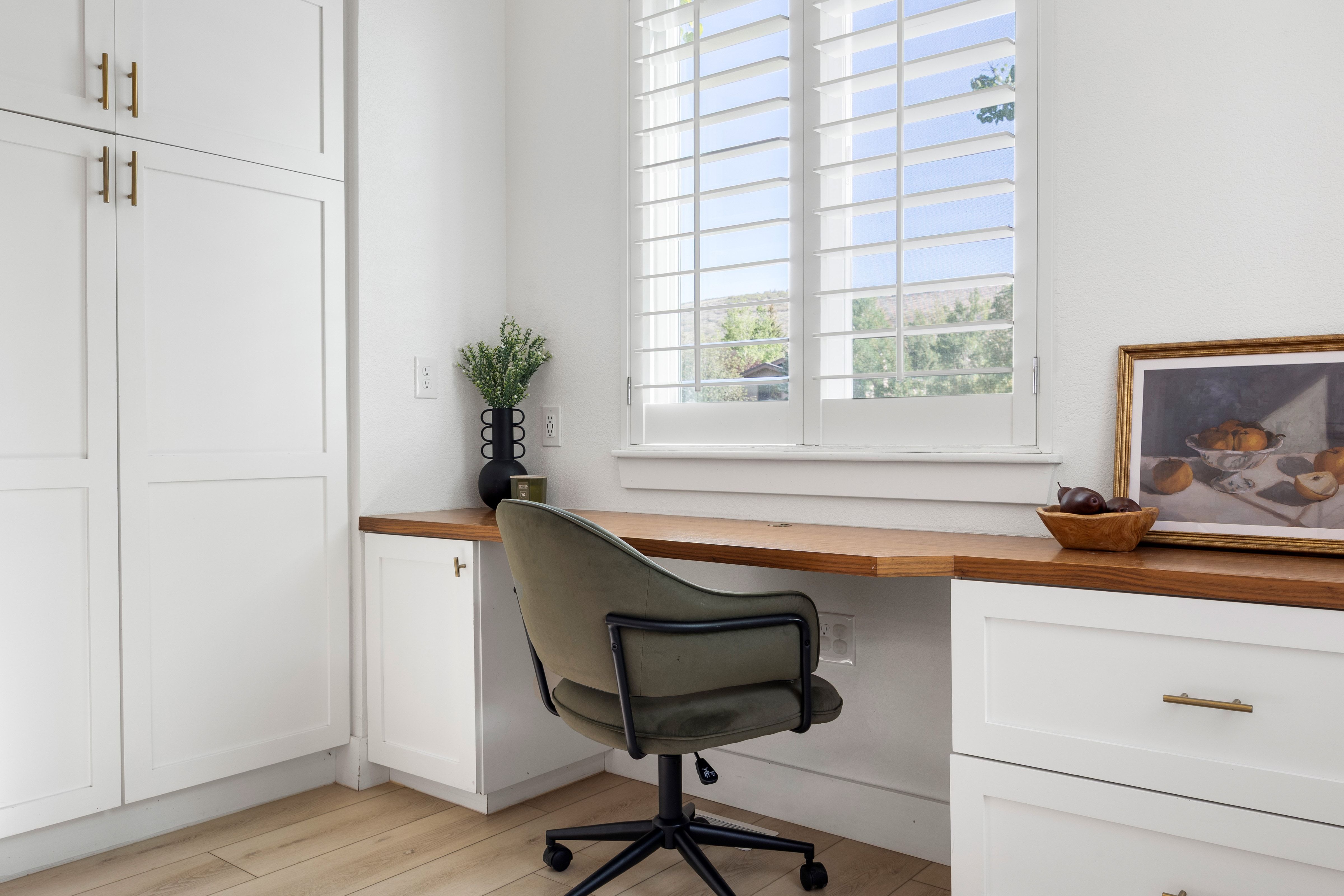 Built-in desk featuring a wood-finish countertop, white cabinetry with gold-tone hardware, and plantation shutters