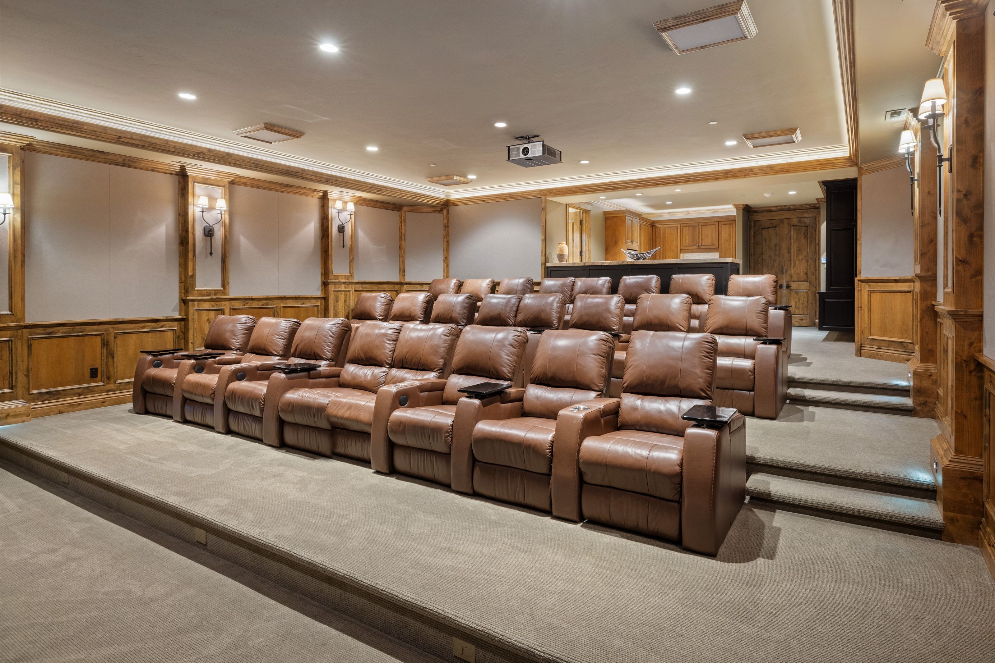 Dedicated home theater featuring tiered seating, built-in projector, recessed lighting, wood-paneled walls, and integrated sconces