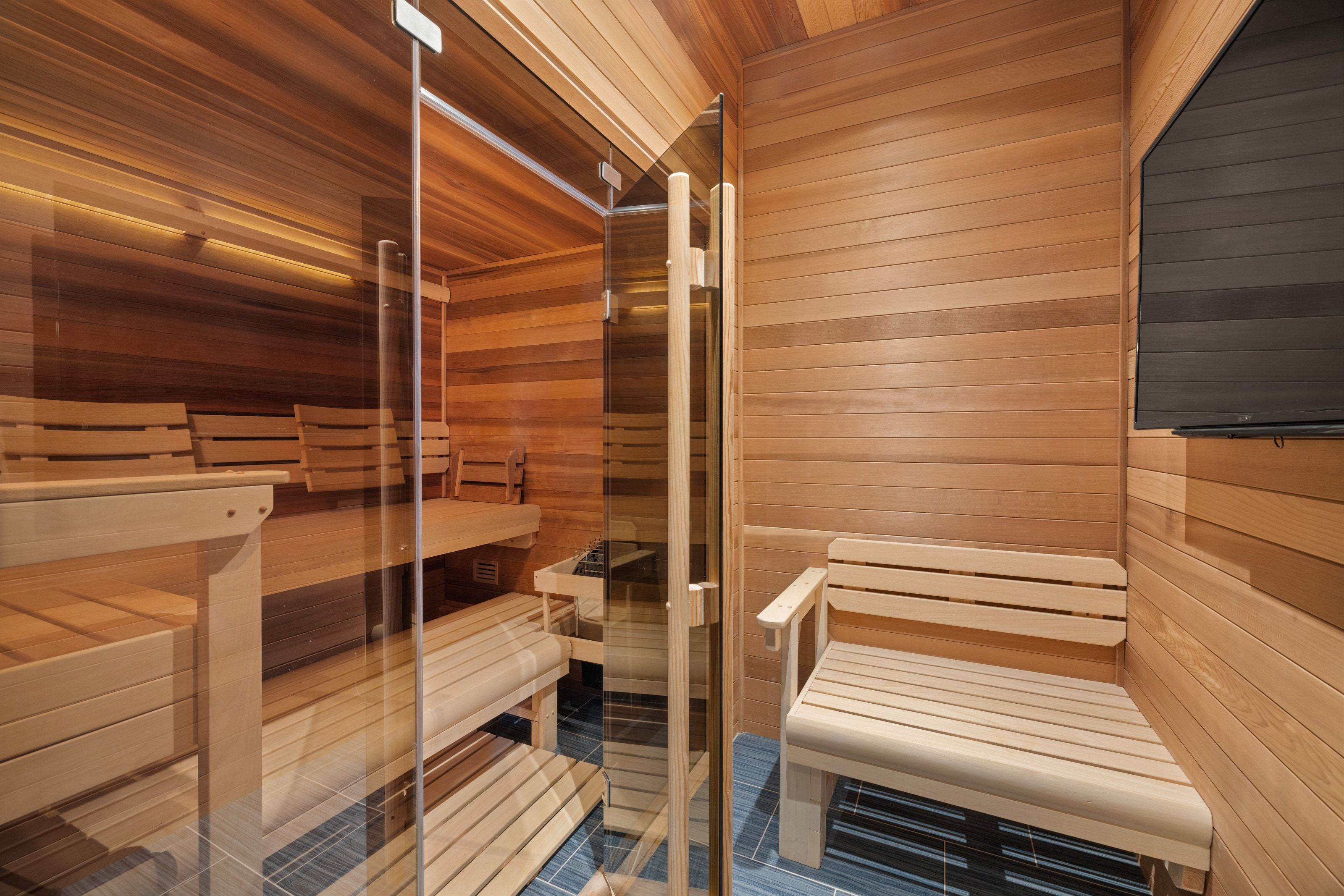 Private sauna featuring clear glass door, multiple slatted wood benches, slatted wood walls and ceiling, and dark tile flooring