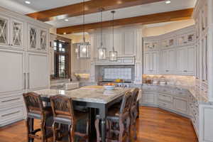 Gourmet kitchen featuring a large central island with seating, extensive custom cabinetry, granite countertops, wood-finish flooring, and exposed ceiling beams