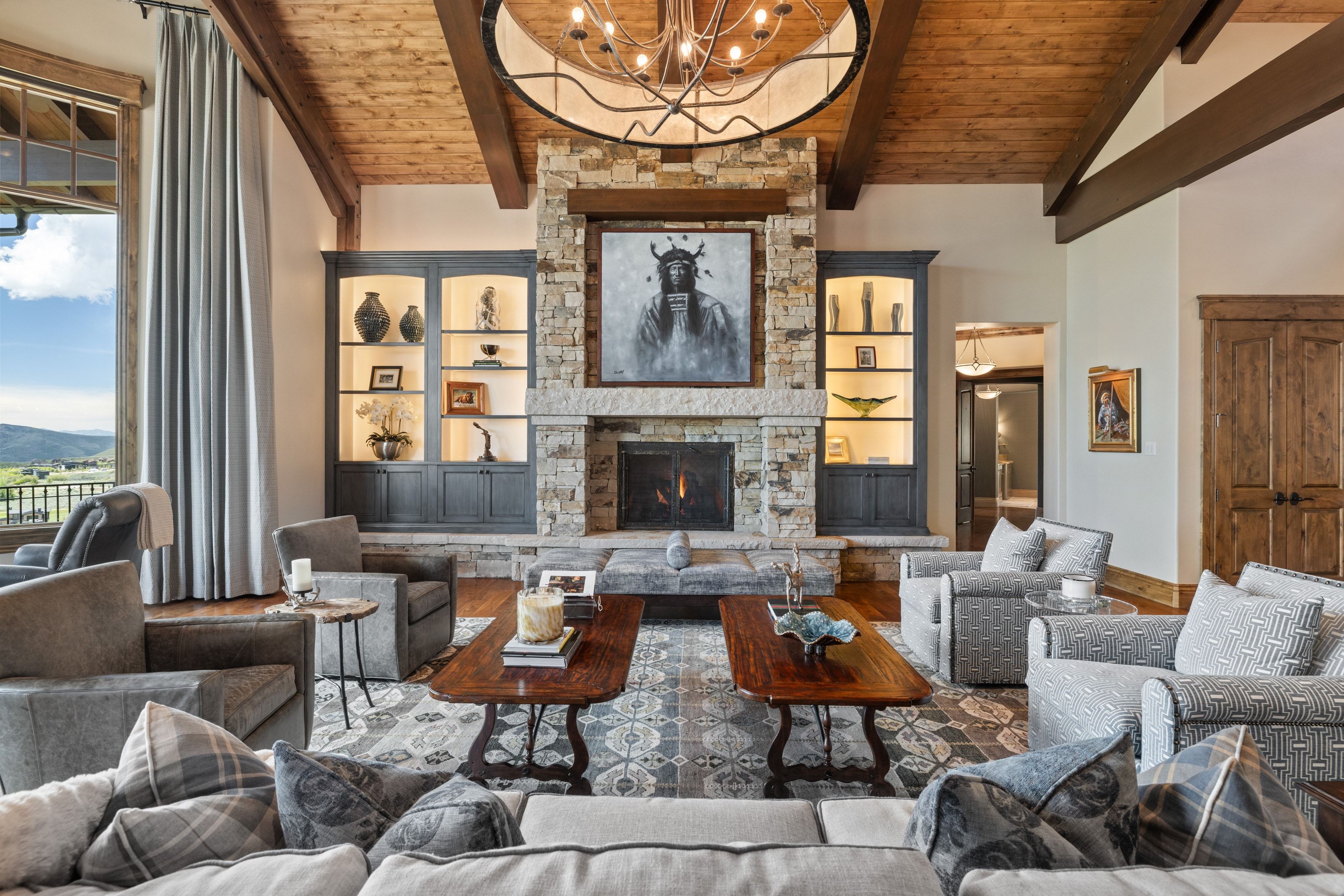 Spacious living area featuring a stone fireplace with a substantial hearth, exposed wood beam ceiling, wood-finish flooring, built-in illuminated display cabinetry, and a large window with a scenic outdoor landscape