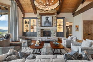 Spacious living area featuring a stone fireplace with a substantial hearth, exposed wood beam ceiling, wood-finish flooring, built-in illuminated display cabinetry, and a large window with a scenic outdoor landscape