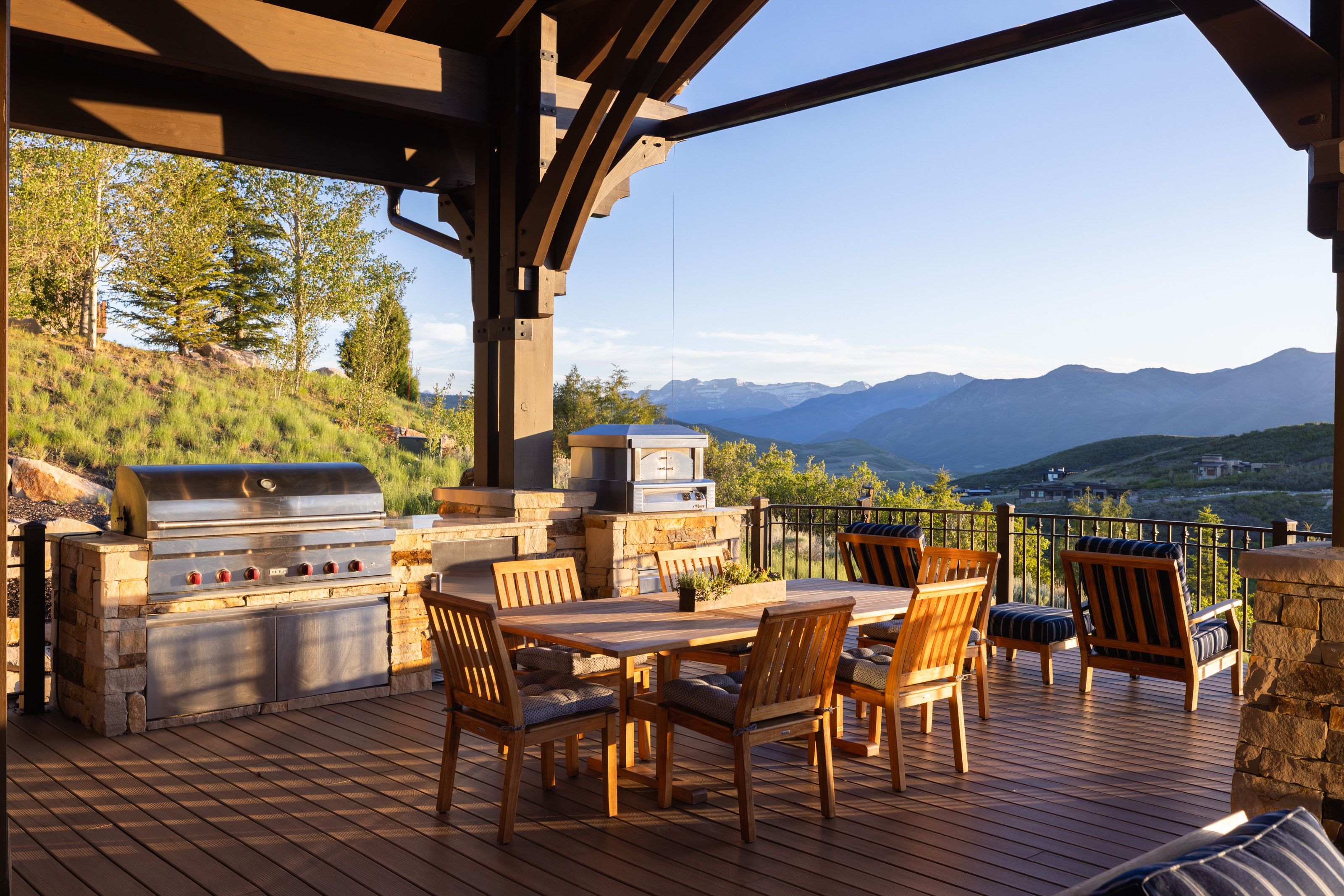 Covered outdoor deck featuring composite decking, exposed timber beams, a built-in outdoor kitchen with stainless steel grill, a stone-clad pizza oven, and a metal railing