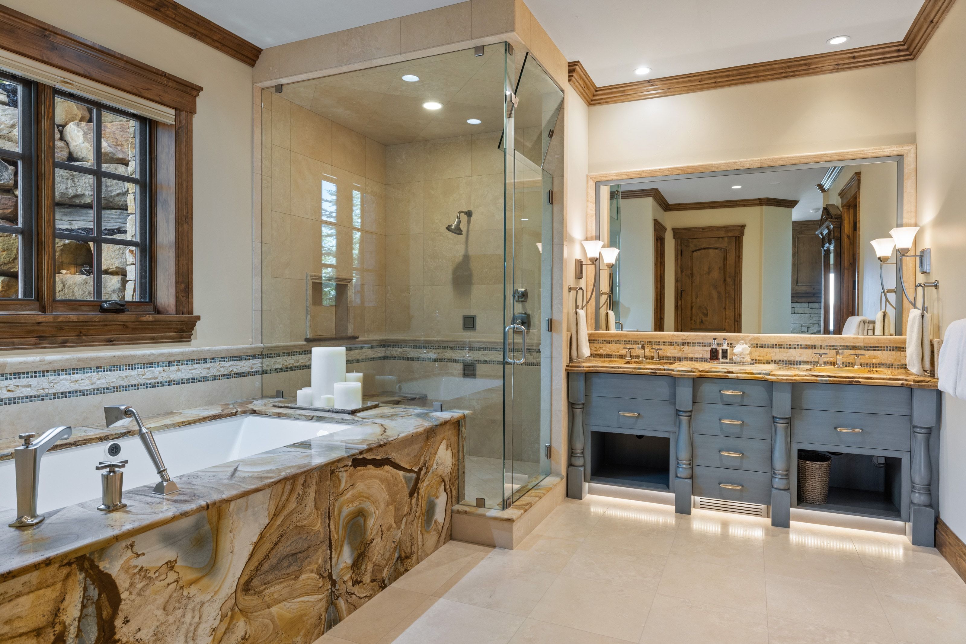 Spacious bathroom featuring a built-in bathtub with a striking natural stone surround, a separate glass-enclosed shower, a double vanity with undermount sinks, and integrated under-cabinet lighting