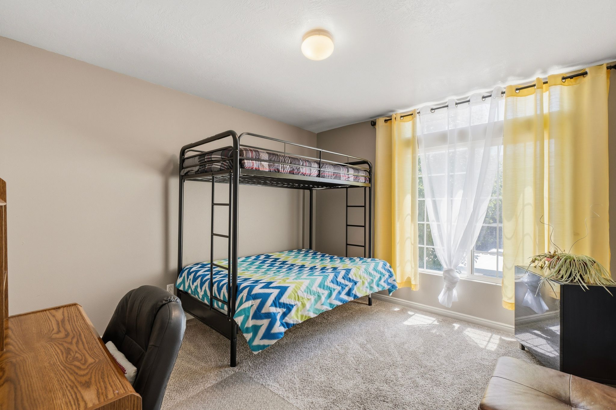 Bedroom with plush carpet flooring, neutral painted walls, and a large window featuring sheer and yellow drapes
