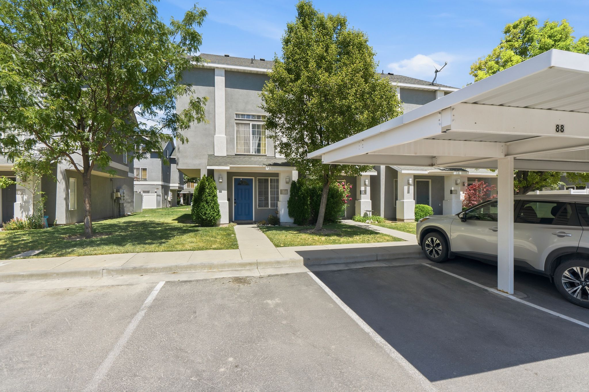 Two-story stucco townhouse with a bright blue entry door, an adjacent covered carport, manicured front landscaping, and mature trees