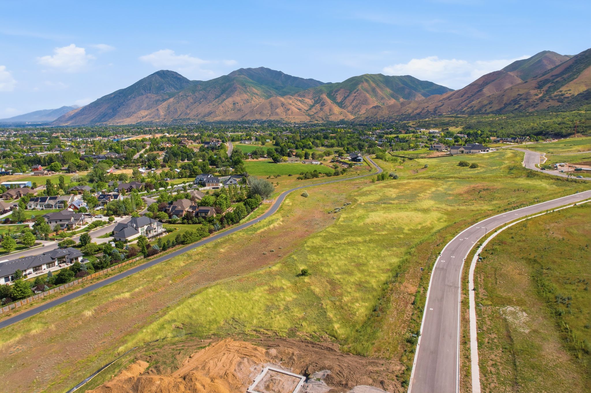 Expansive undeveloped land featuring a paved road with an integrated sidewalk, surrounding residential structures, and distant mountains under a clear sky