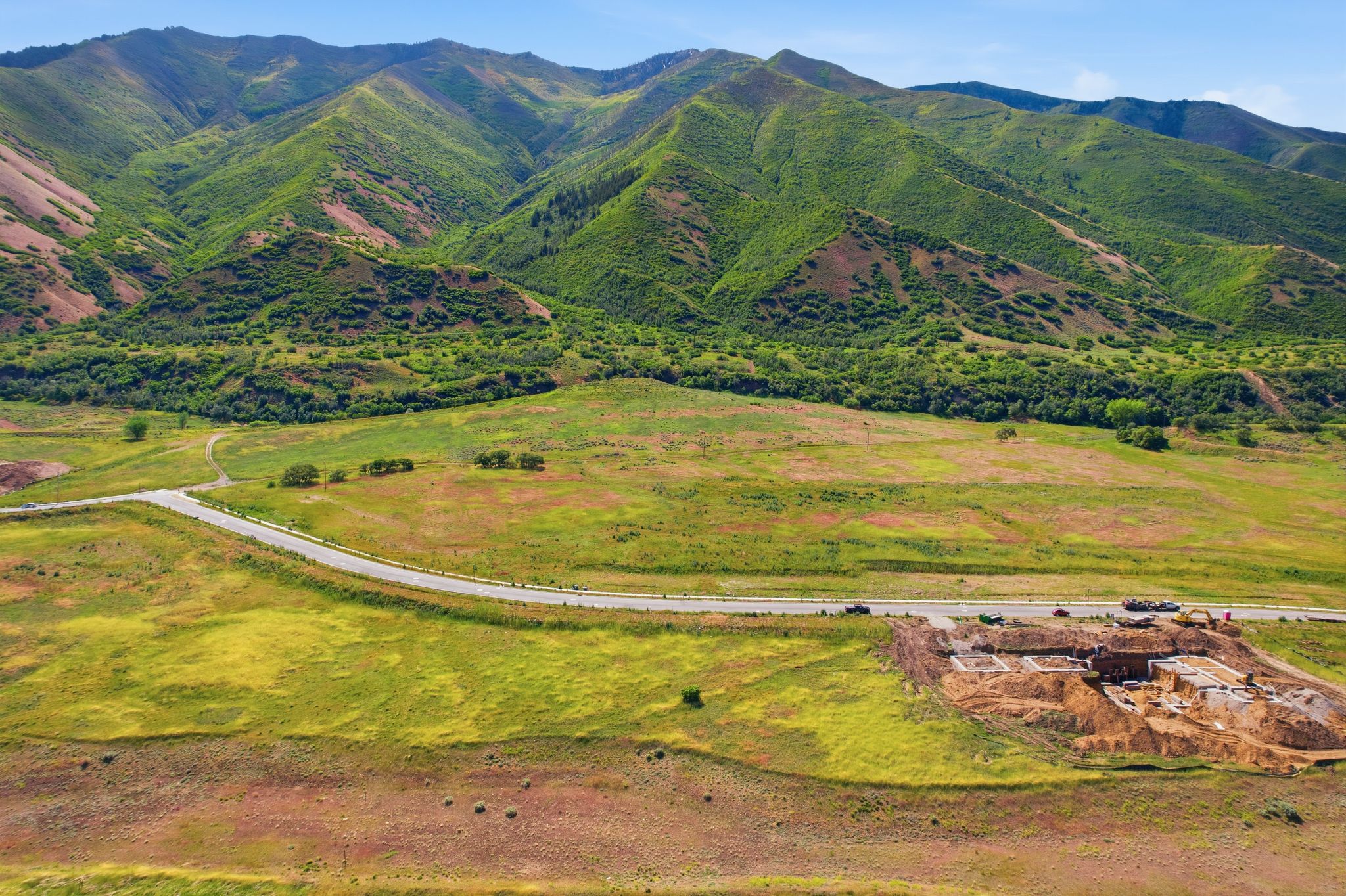 Expansive undeveloped terrain featuring a winding paved road, lush green mountains, and a clear blue sky