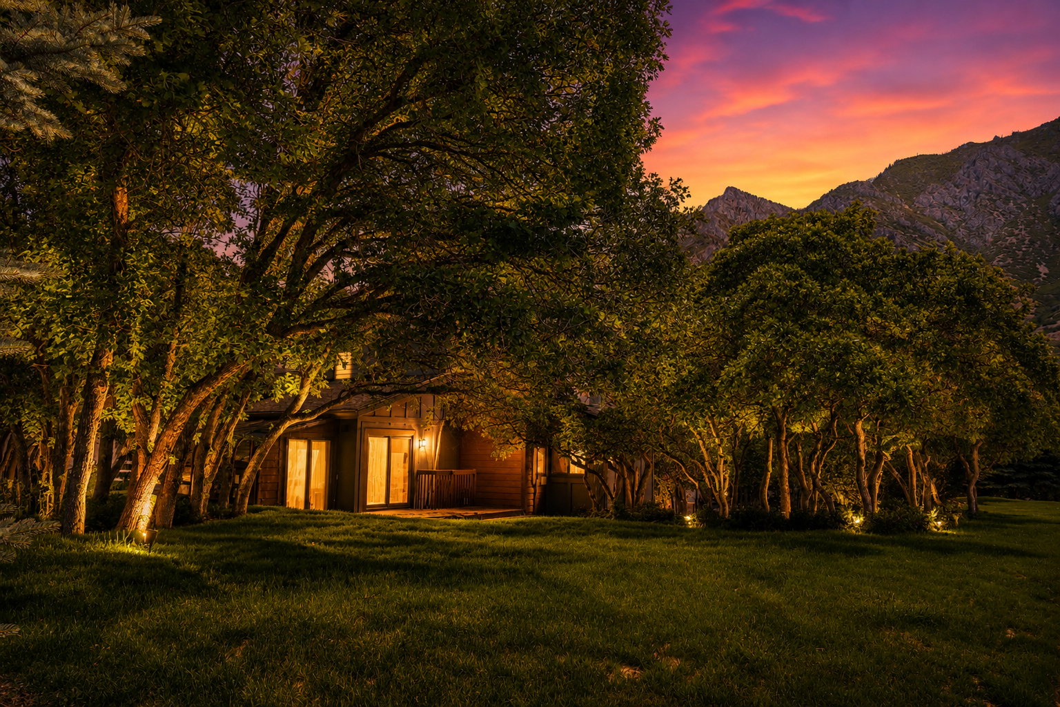 Exterior featuring a mountainous backdrop, mature trees with uplighting, a manicured lawn, and a private patio with exterior lighting