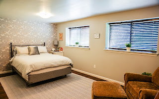 Bedroom featuring a patterned accent wall, two distinct windows with blinds, dark wood-finish flooring, and a flush-mount ceiling light