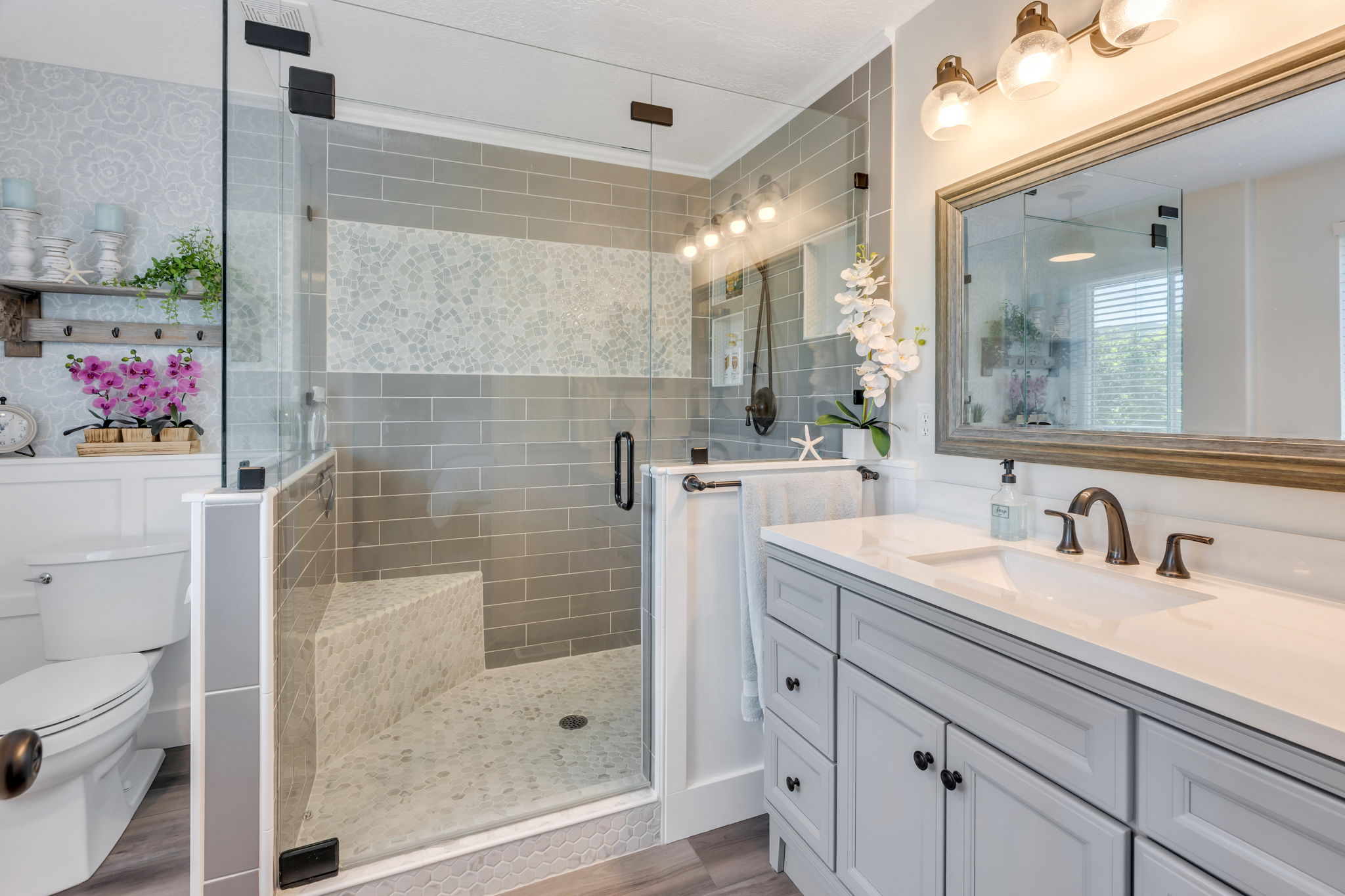 Contemporary bathroom featuring a spacious glass-enclosed shower with subway tile and mosaic accents, a built-in shower bench, a single vanity with light gray cabinetry and a white countertop, and a large framed mirror with overhead lighting